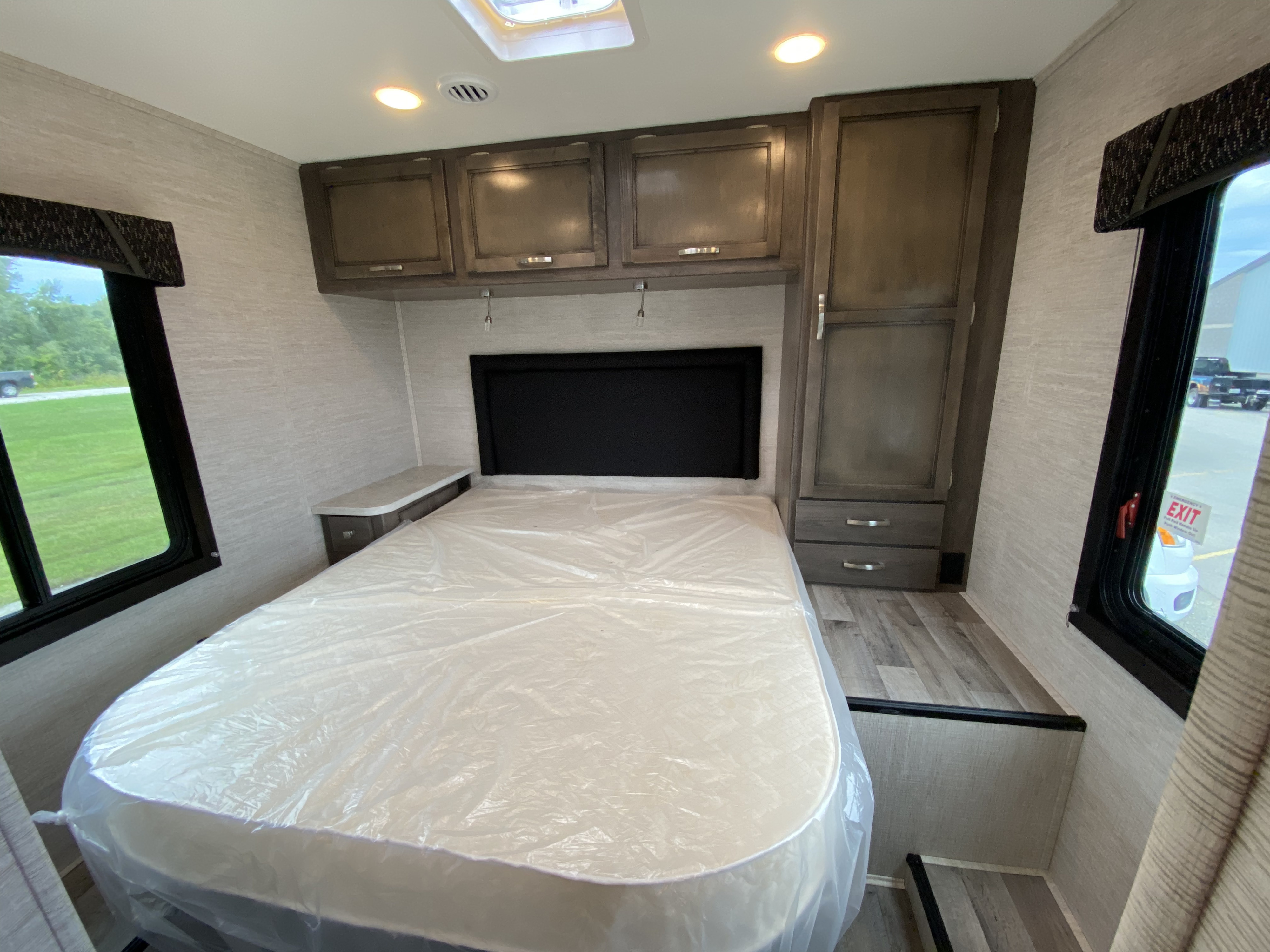 This RV rental bedroom features a new bed with plastic cover, wood-look cabinets, side nightstand, windows, and light textured walls.. Jayco Redhawk 27N 2022