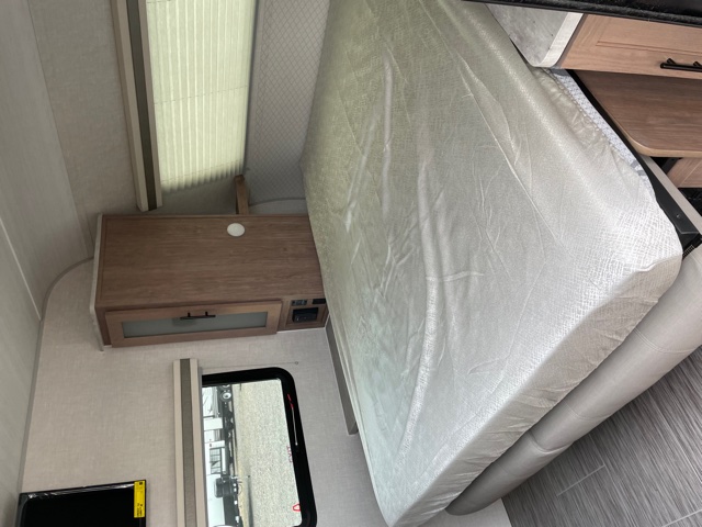 View inside an RV rental: a spacious bed lifted for access, a wooden nightstand with storage, and a window with blinds.. Forest River No Boundaries 2021