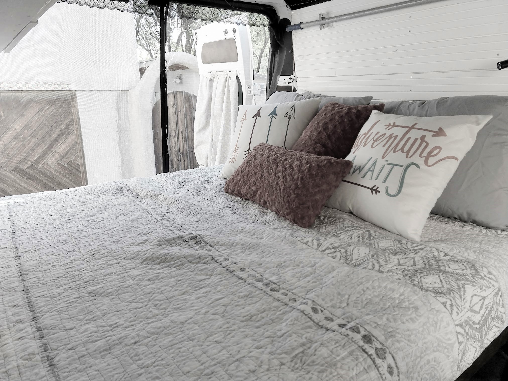 Inside an RV rental: a made bed with an "Adventure Awaits" pillow, white paneled walls, and open back doors.. Dodge Promaster 2500 High Roof 2020
