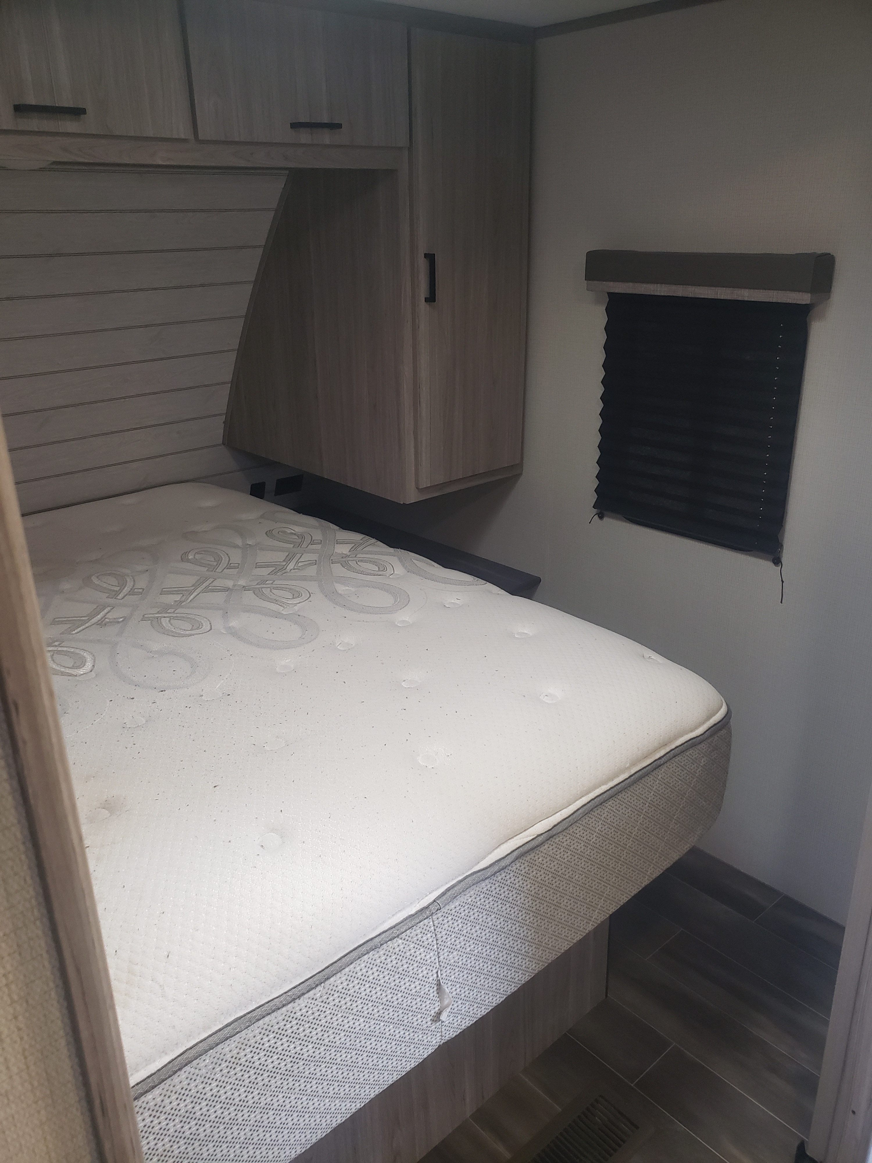 Inside an RV rental: Bedroom area with a white mattress (some stains), wood cabinets, and a black window blind.. Keystone Passport 2021