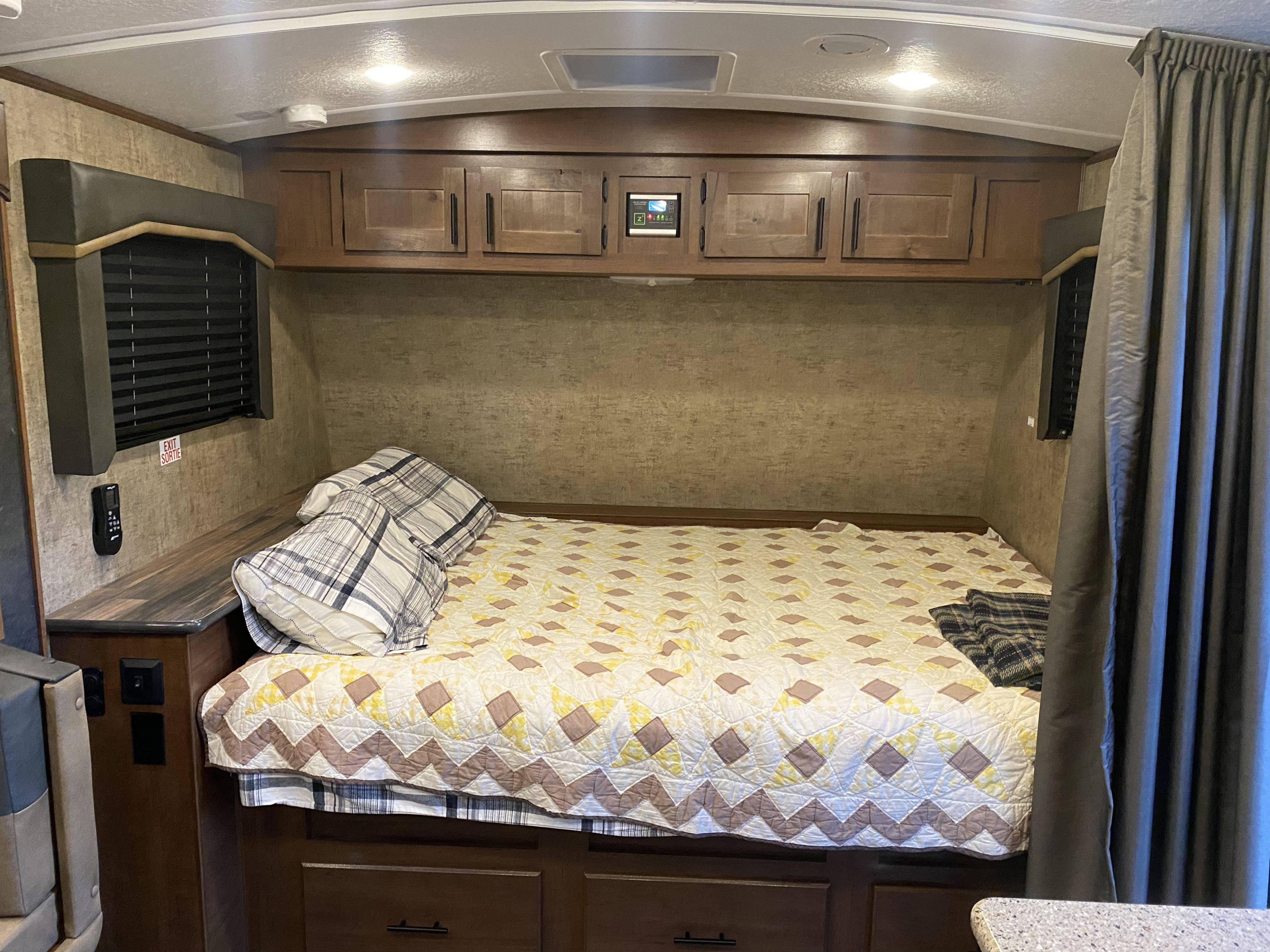 RV rental bedroom: a cozy bed with quilt & pillows, flanked by cabinets & window, with an overhead control panel.. Outdoors Rv Manufacturing Creek Side 2021