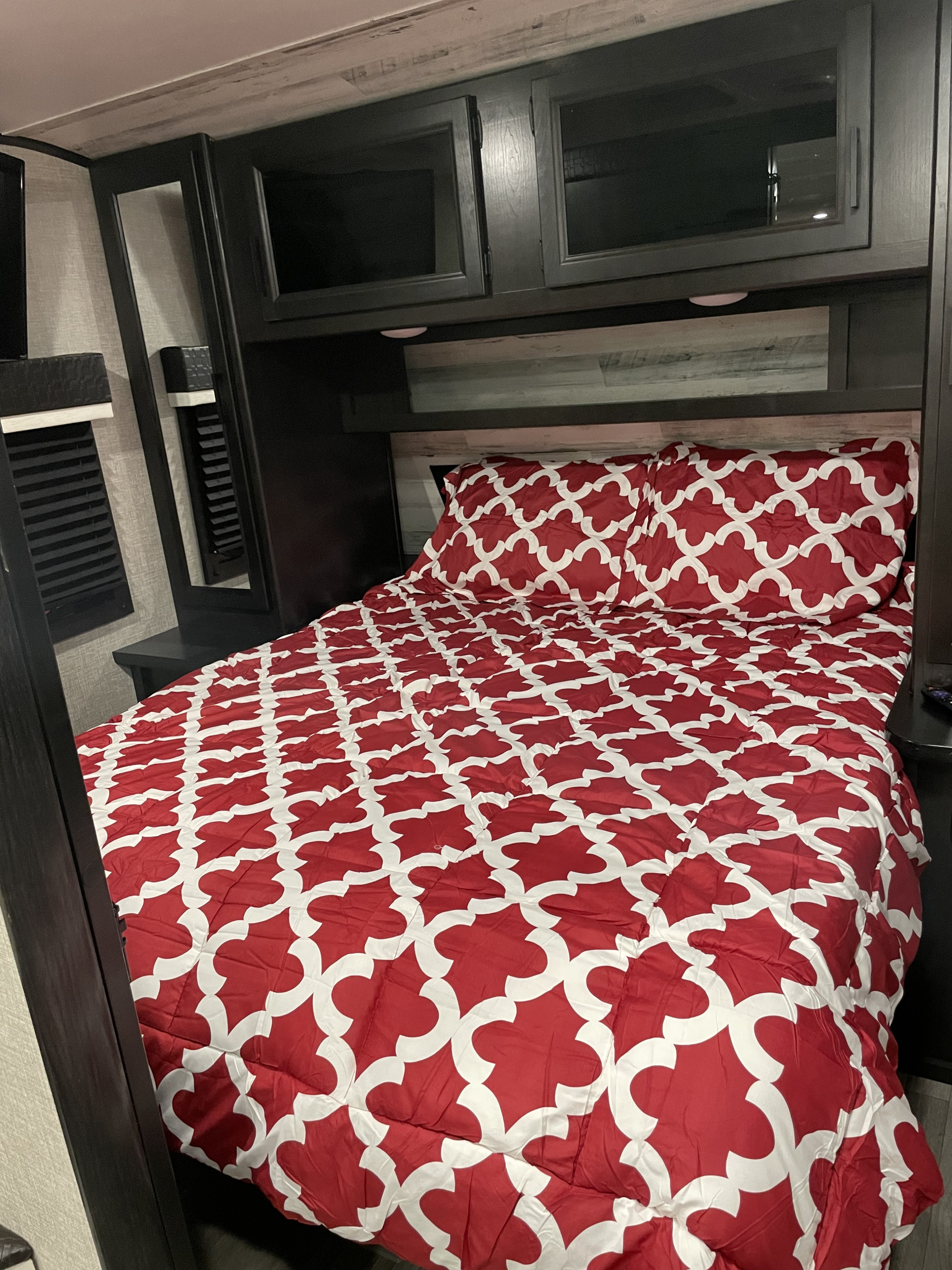 Inside a comfortable RV rental bedroom, featuring a vibrant red and white patterned bed, dark wood cabinets, and shelving.. Jayco Jay Feather 2022
