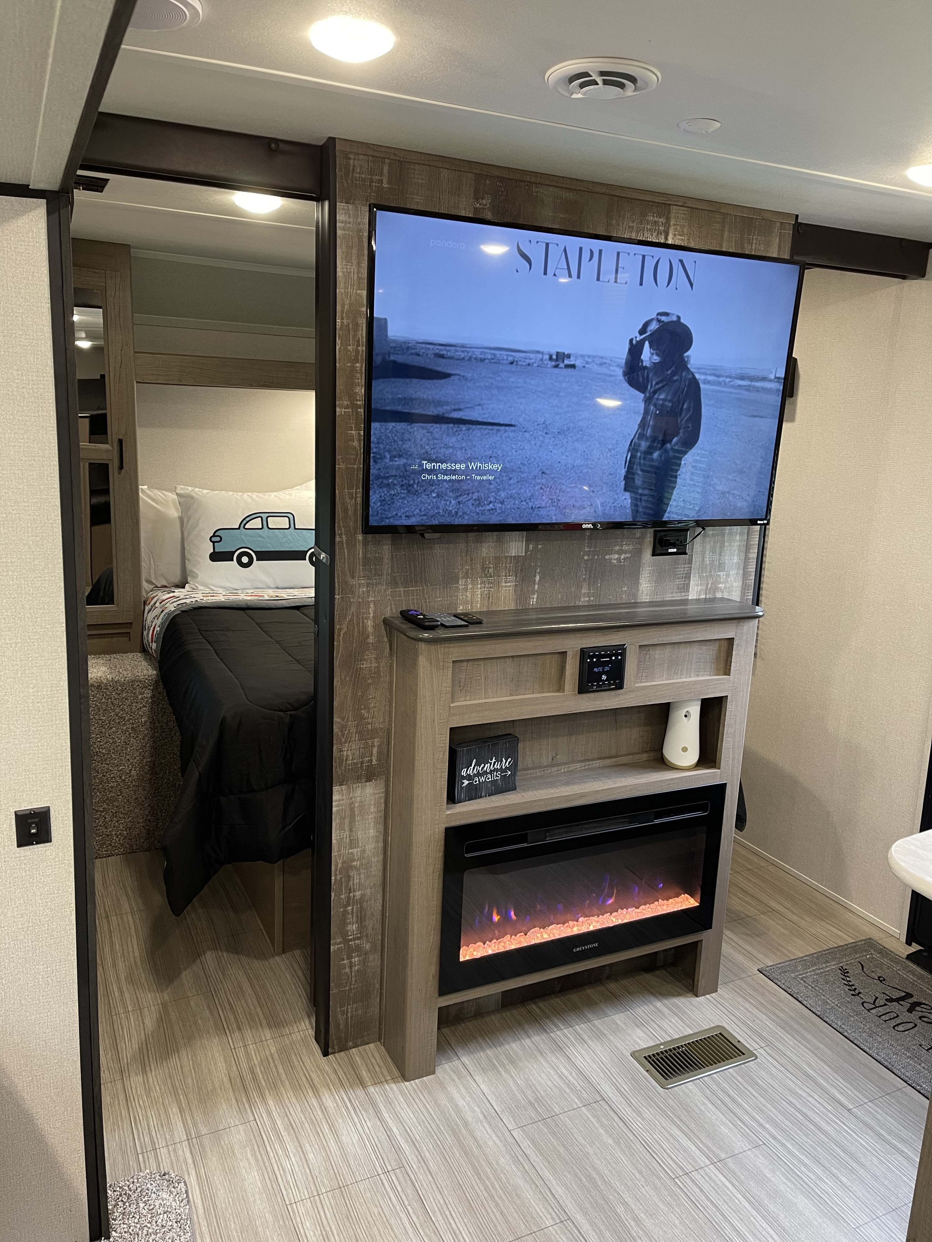 Cozy RV rental interior: TV playing Chris Stapleton, electric fireplace, and a peek into a comfy bedroom with a car pillow.. Keystone Hideout 2021