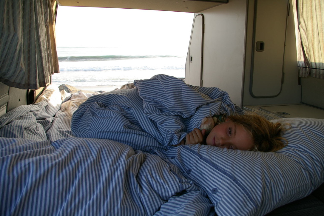 RV rental comfort: A child rests in striped bedding, observing the ocean waves from inside a camper van.. Volkswagen Westfalia 1988