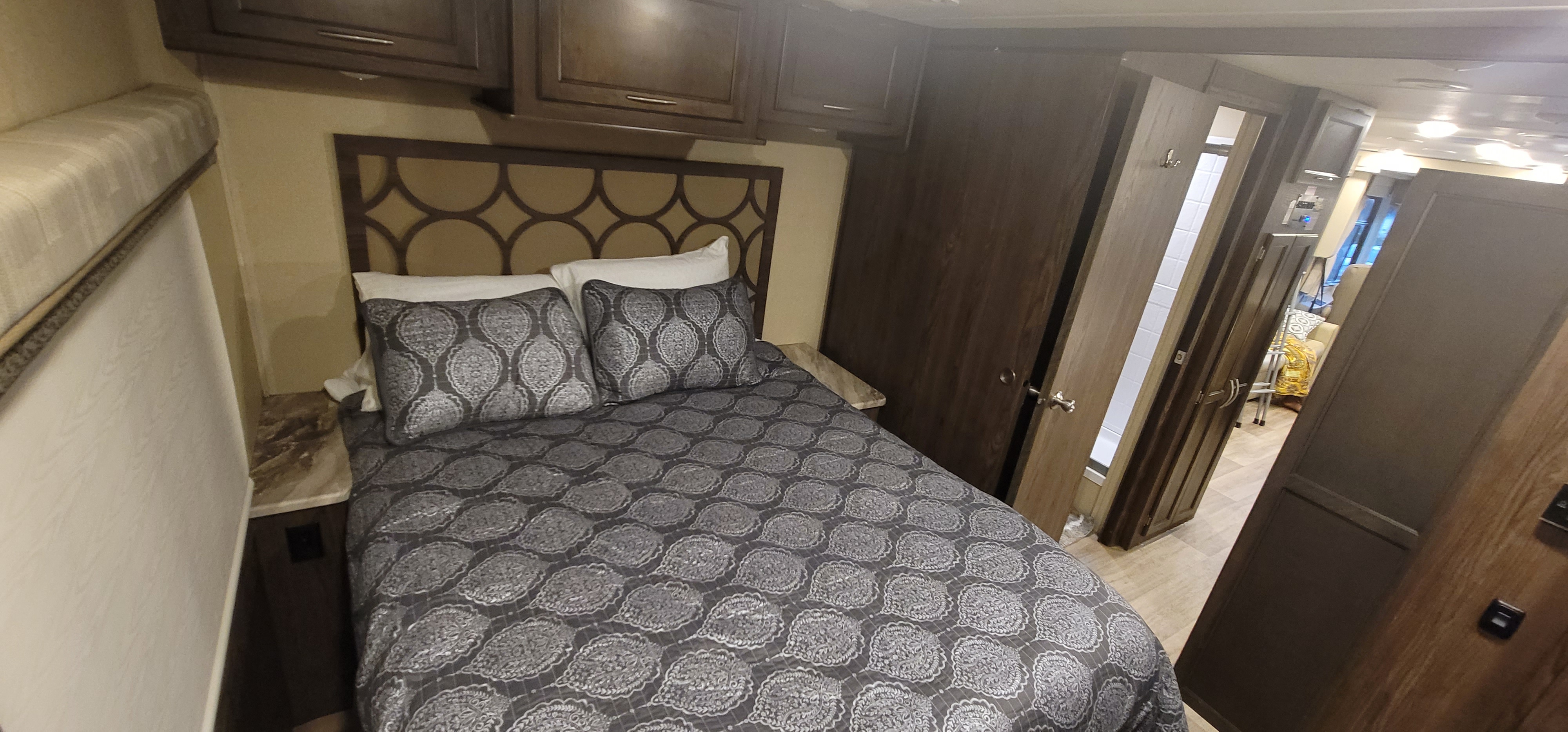 A clean and inviting RV rental bedroom. Features a comfortable bed with patterned covers, overhead cabinets, and a side door.. Coachmen Pursuit 2019