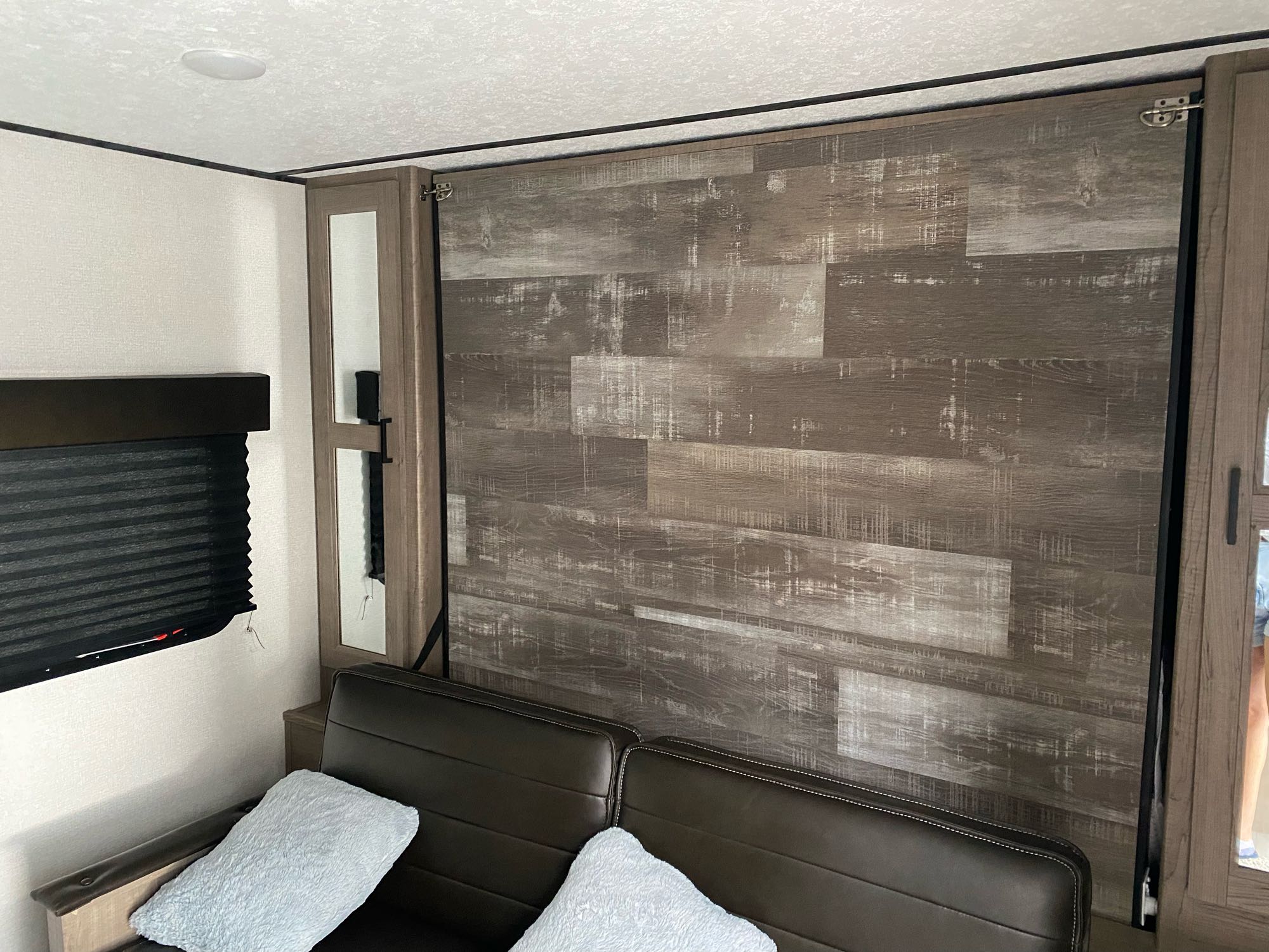 Inside an RV rental: a wood-paneled Murphy bed wall above a brown sofa with light blue pillows, beside a window.. Keystone Hideout 2021