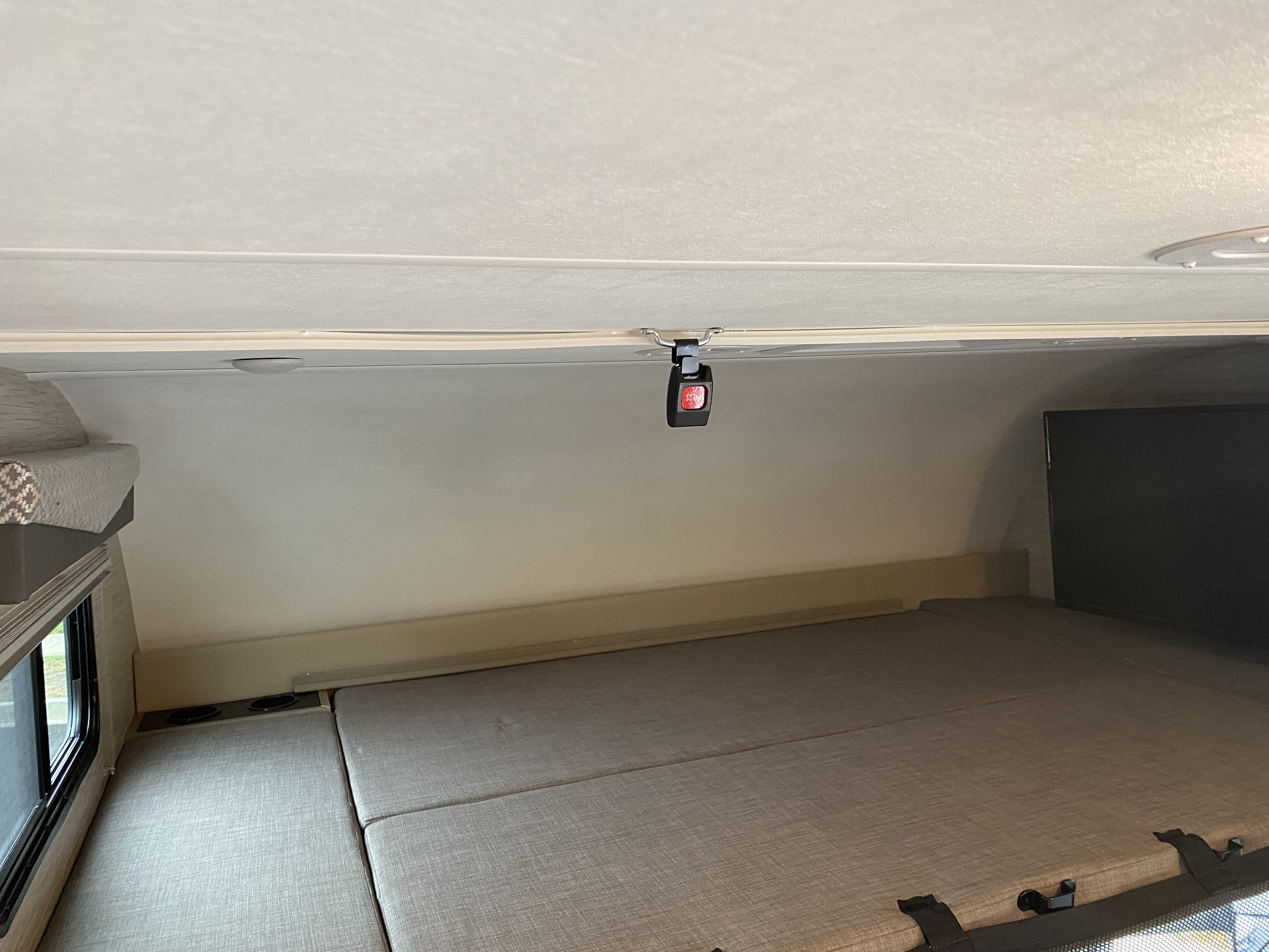 Inside an RV rental: a cab-over bunk with a TV, window, comfortable bedding, and a hanging safety buckle.. Thor Four Winds 2022