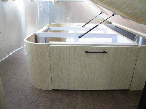 Modern RV rental interior with lift-up bed storage unit. Features light wood, curved design, and sleek handle for accessible storage.. Airstream International 2016