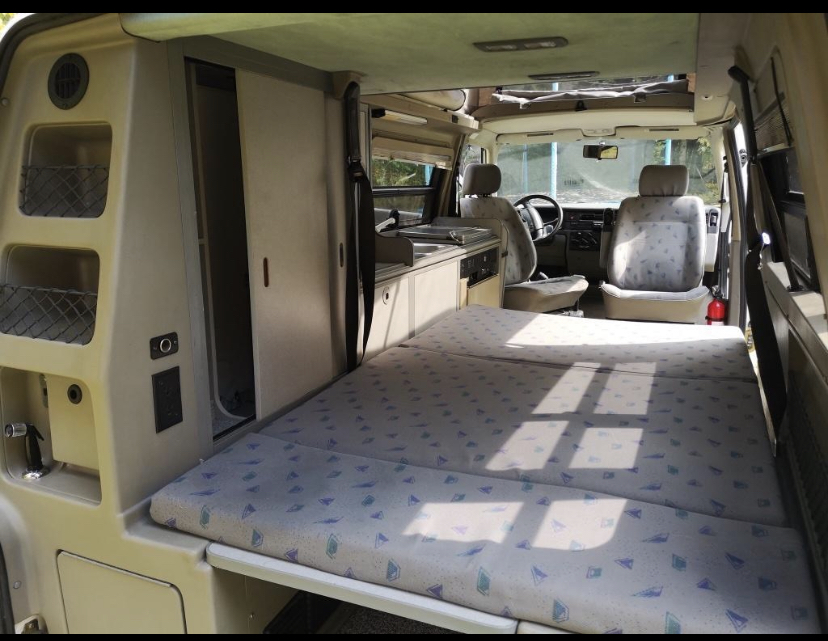 Inside an RV rental: a light-colored camper van with a fully extended patterned fold-out bed, compact kitchenette, and front seats.. Winnebago Eurovan Camper Eurovan Camper 1997