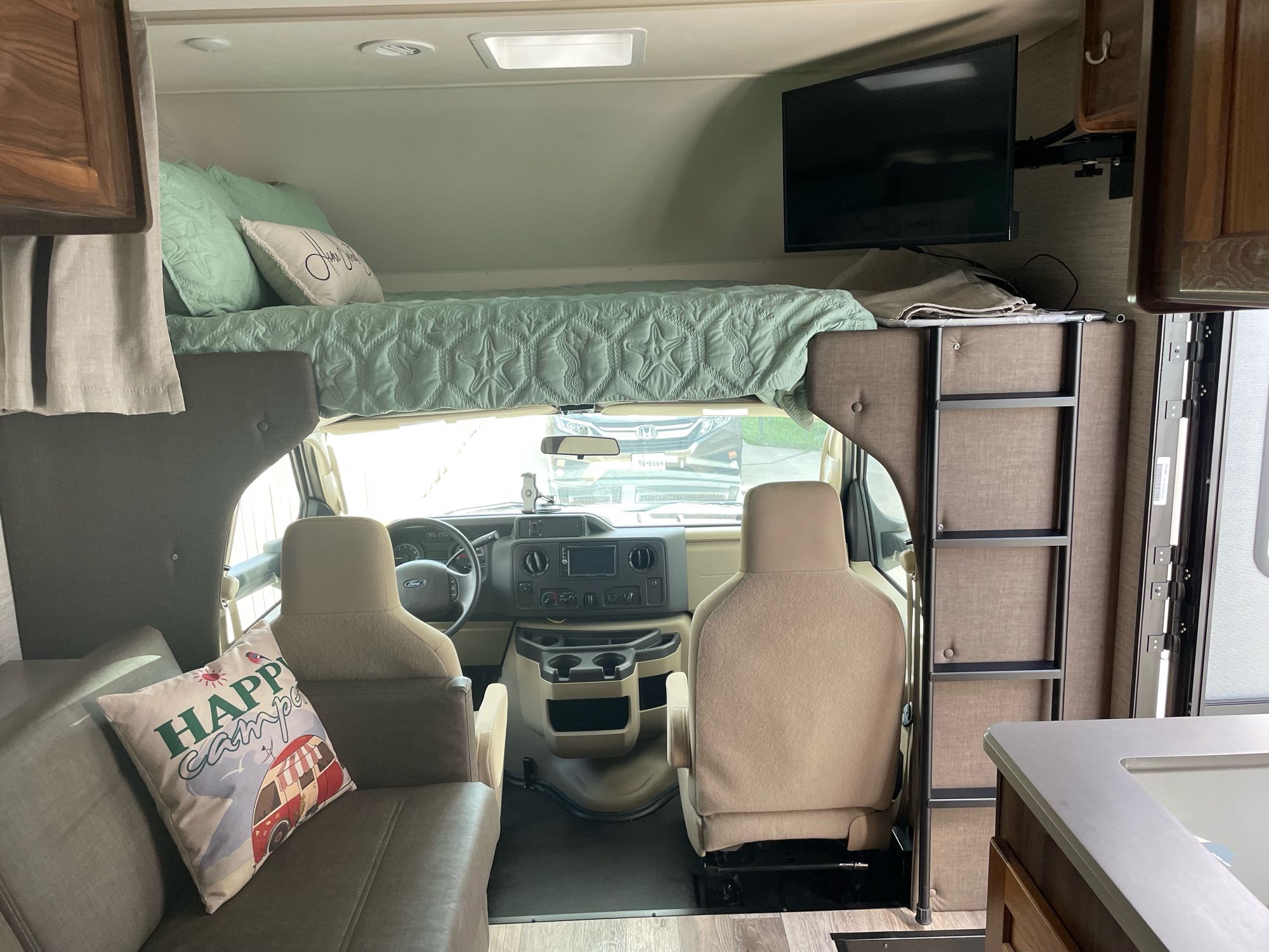 Inside this **rv rental**, a large bed is above the driver's area, with a sofa and TV in the living space. Driver's cabin visible.. Winnebago Outlook 2020