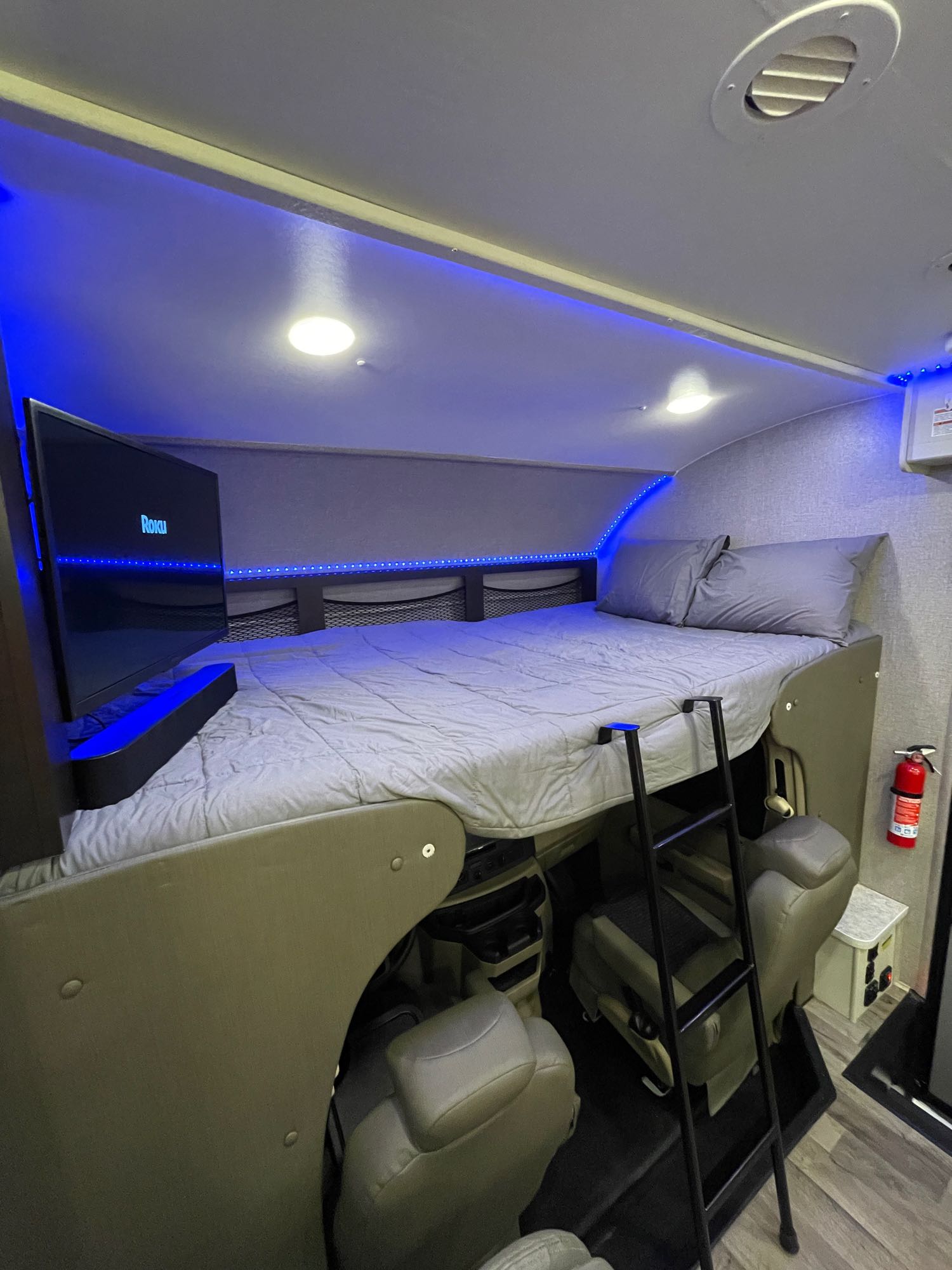 Cozy rv rental cab-over bunk bed with TV, blue LED lighting, and ladder. Features grey bedding above front seats.. Jayco Redhawk 2021