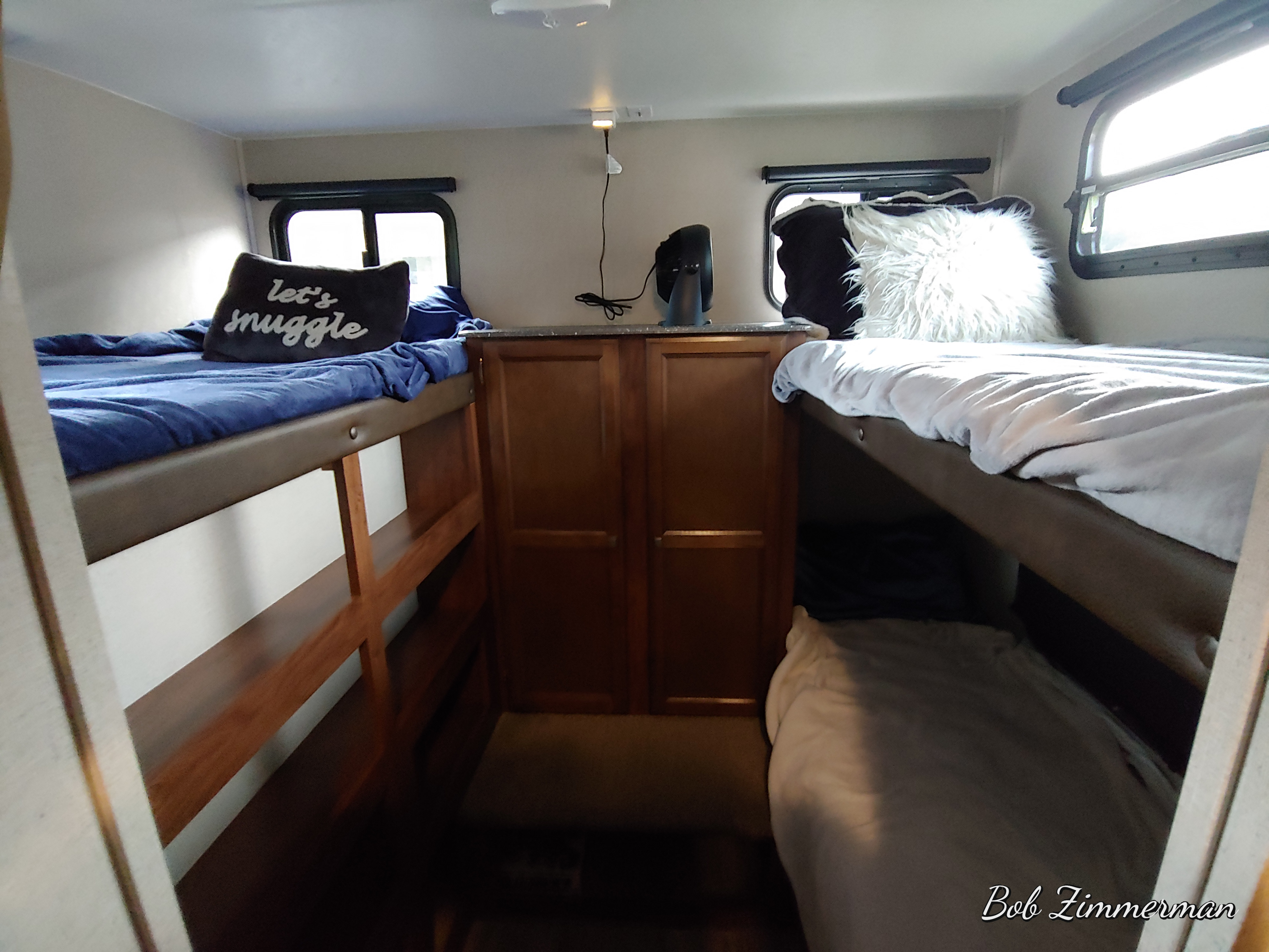 An RV rental interior with bunk beds: blue bedding on left, white on right, plush pillows, and central wood cabinets.. Coachmen Apex 2018