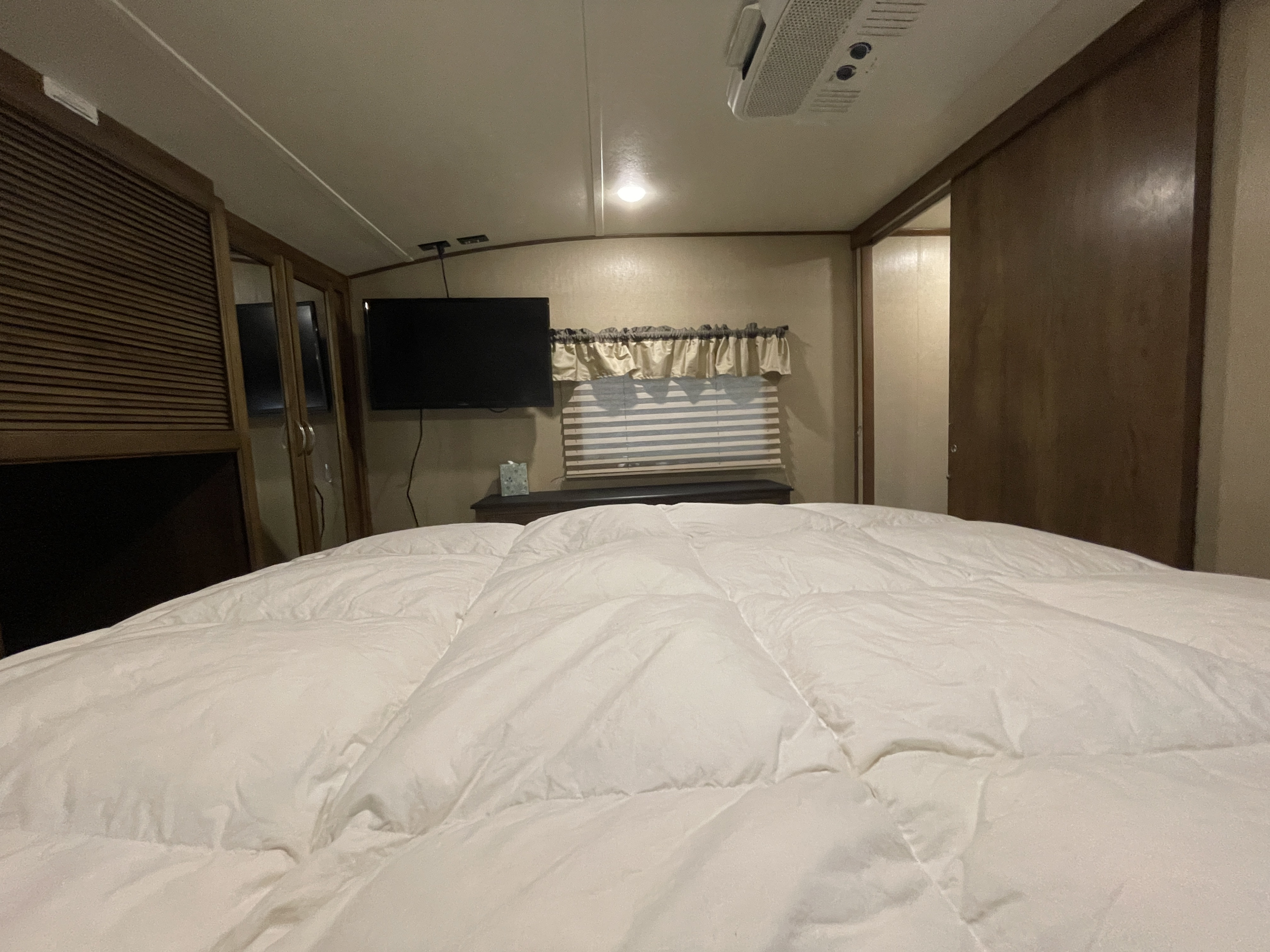 This RV rental bedroom features a comfy white duvet, wall-mounted TV, window with blinds, and ceiling AC unit.. Forest River Coachmen Chaparral 2017