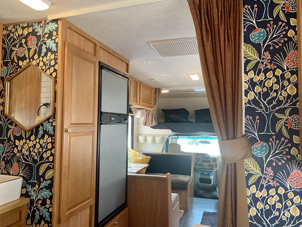 Interior of an RV rental showing custom botanical wallpaper, wood cabinets, fridge, and a cozy living area with bunk.. Winnebago Chalet 2010