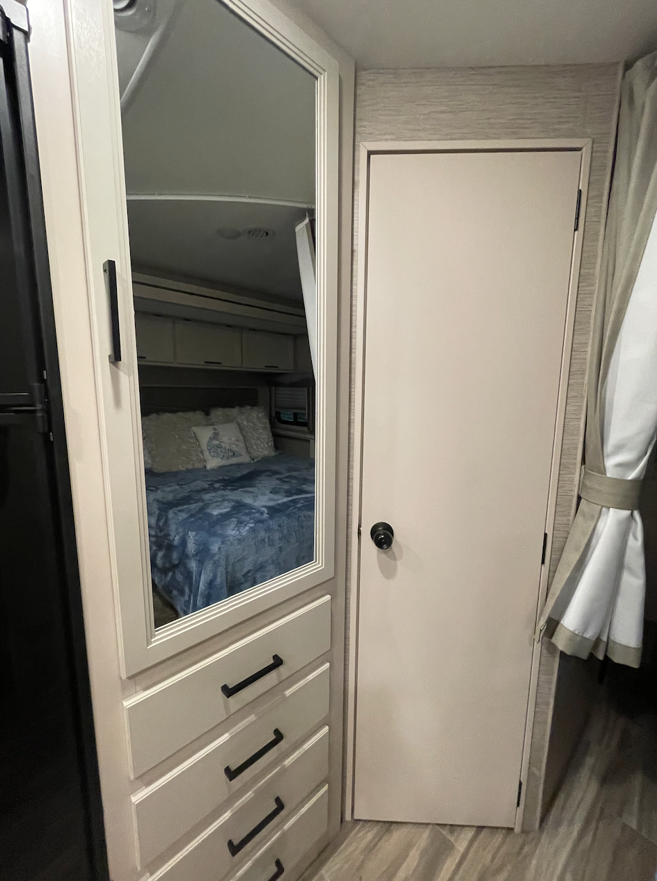 This RV rental features a compact bedroom with a mirrored wardrobe and drawers, reflecting a blue bed and overhead cabinets.. Thor Motor Coach Freedom Elite 2022