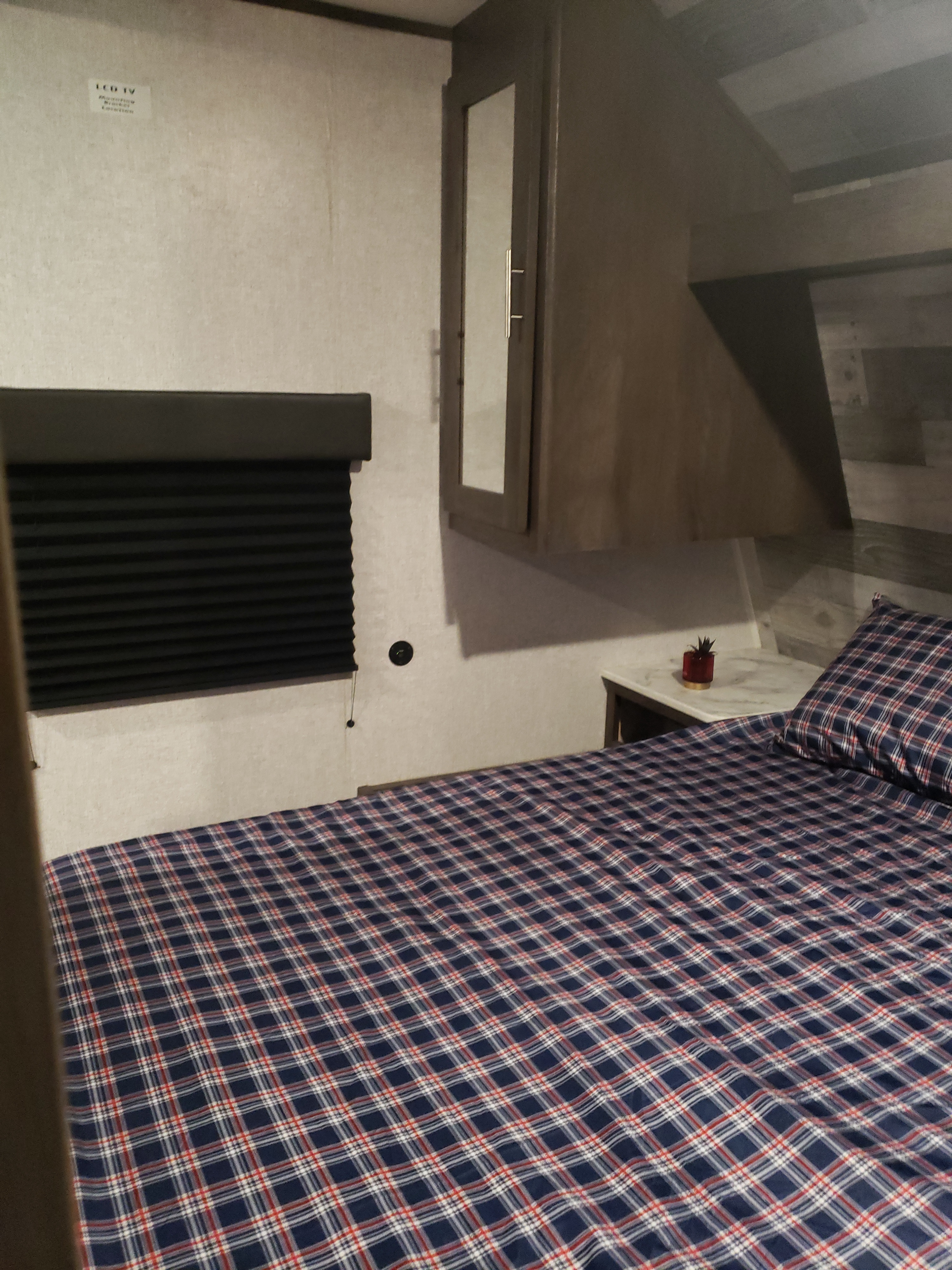 Inside an RV rental bedroom: a bed with blue plaid bedding, overhead cabinets & a small nightstand. Ready for your adventure.. Dutchmen Coleman 2021