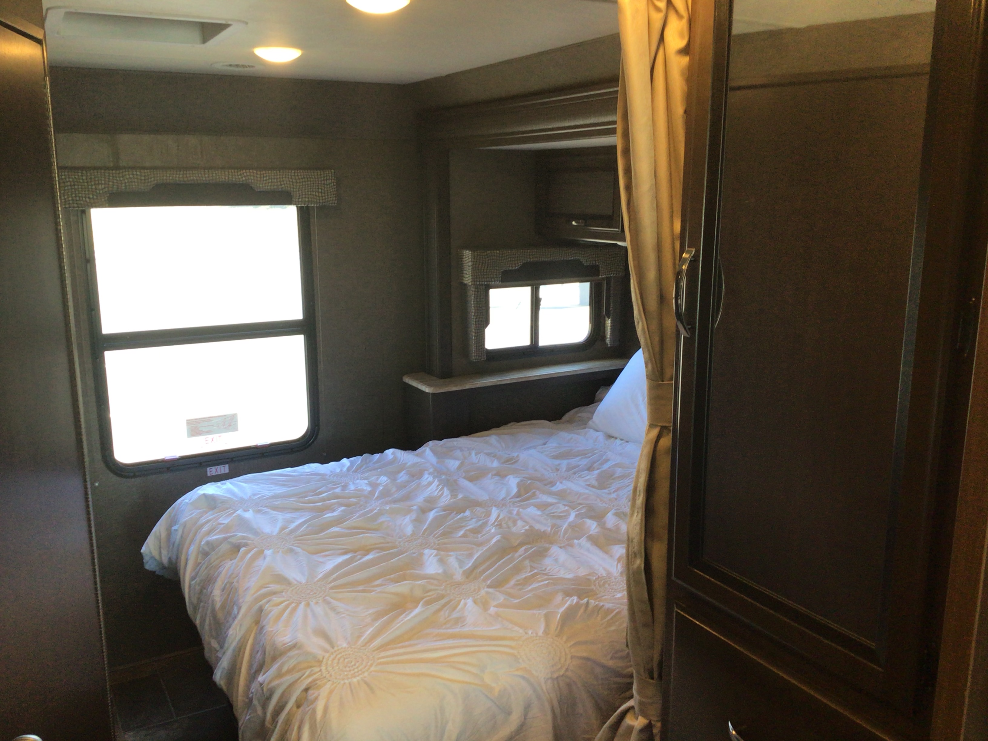 Cozy RV rental bedroom featuring a white bed, bright windows with "EXIT" signs, and dark wood cabinetry.. Thor Motor Coach Vegas 2017