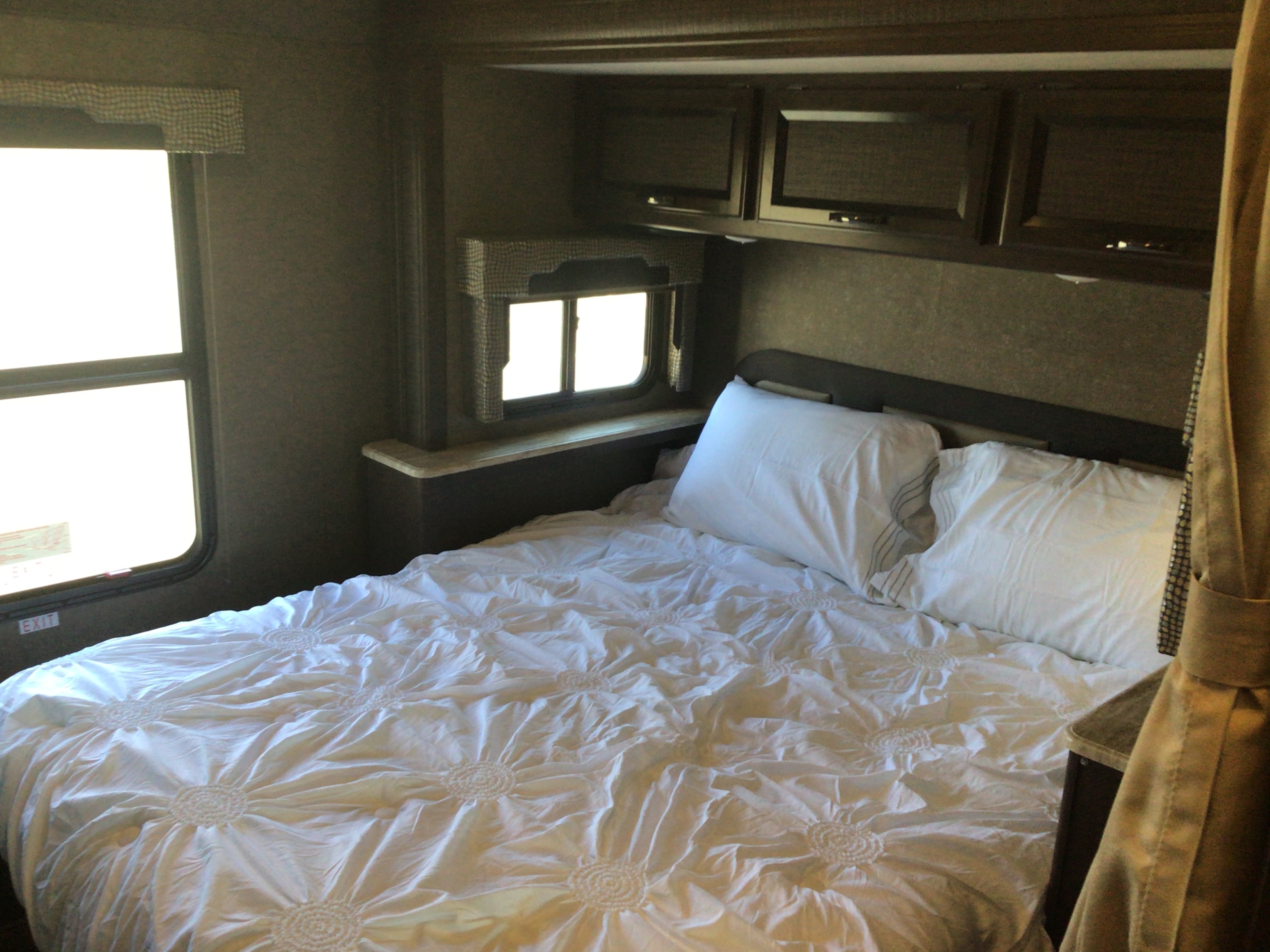 Inside an RV rental: a comfortable bedroom with a white bed, bright windows, and ample overhead storage cabinets.. Thor Motor Coach Vegas 2017