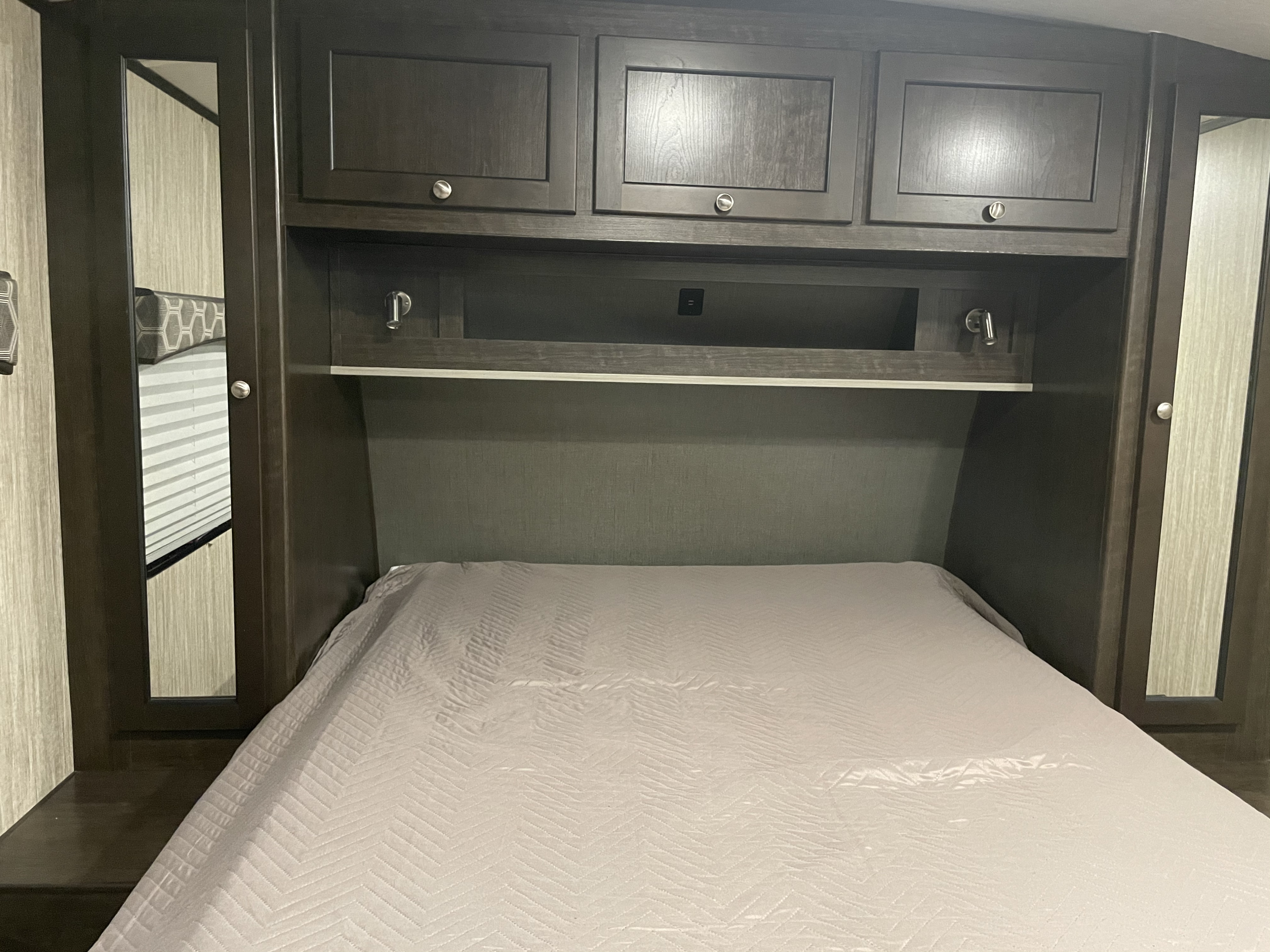 RV rental bedroom with bed, dark overhead cabinets, shelf with USB, reading lights, and mirrored wardrobe doors. Compact mobile comfort.. Cruiser Rv Corp Shadow Cruiser 2020