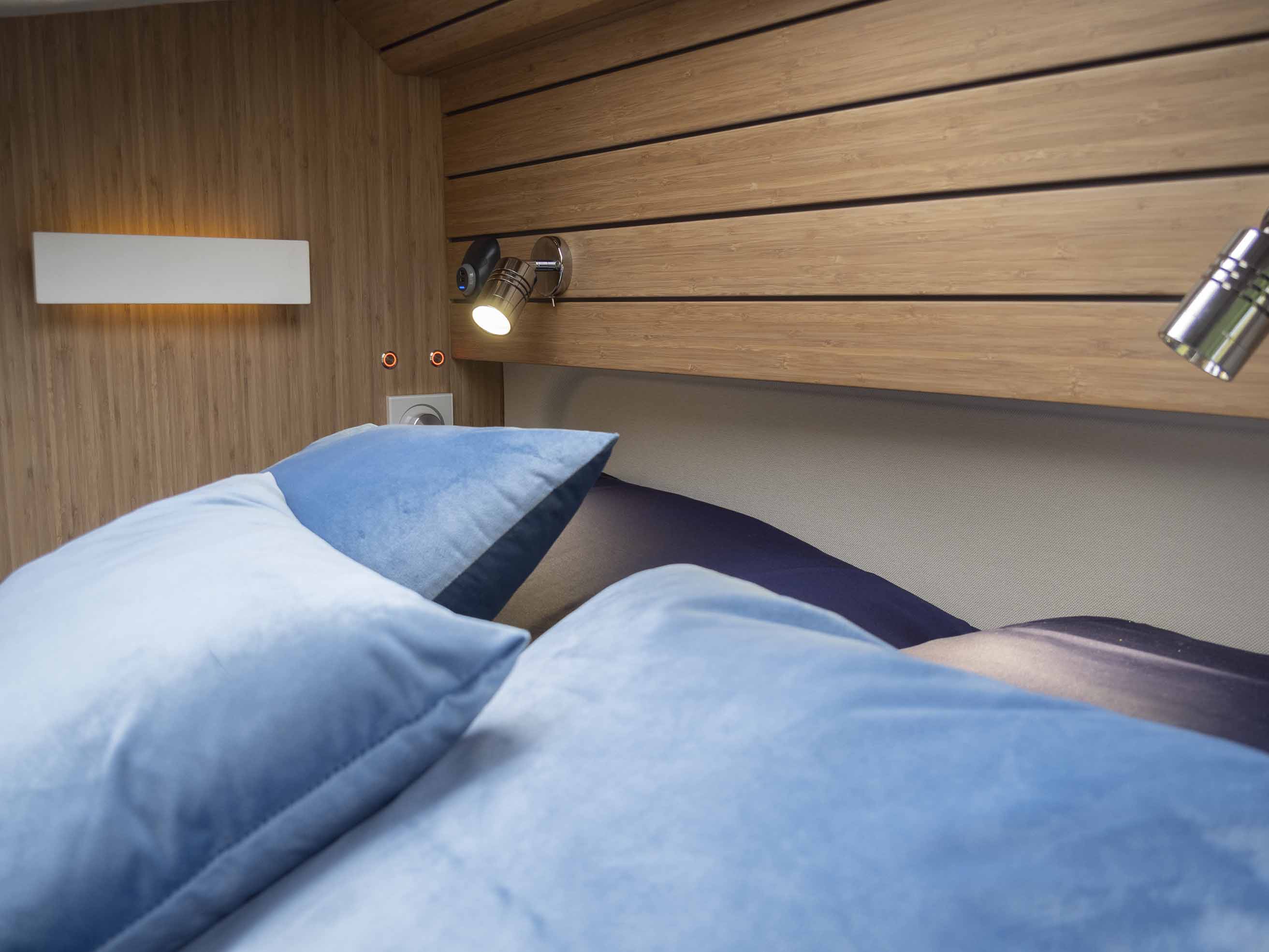 Comfortable RV rental bedroom: plush blue pillows, warm wood paneling, and adjustable reading lights. Ready for travel.. Fiat Ducato MH2 2021