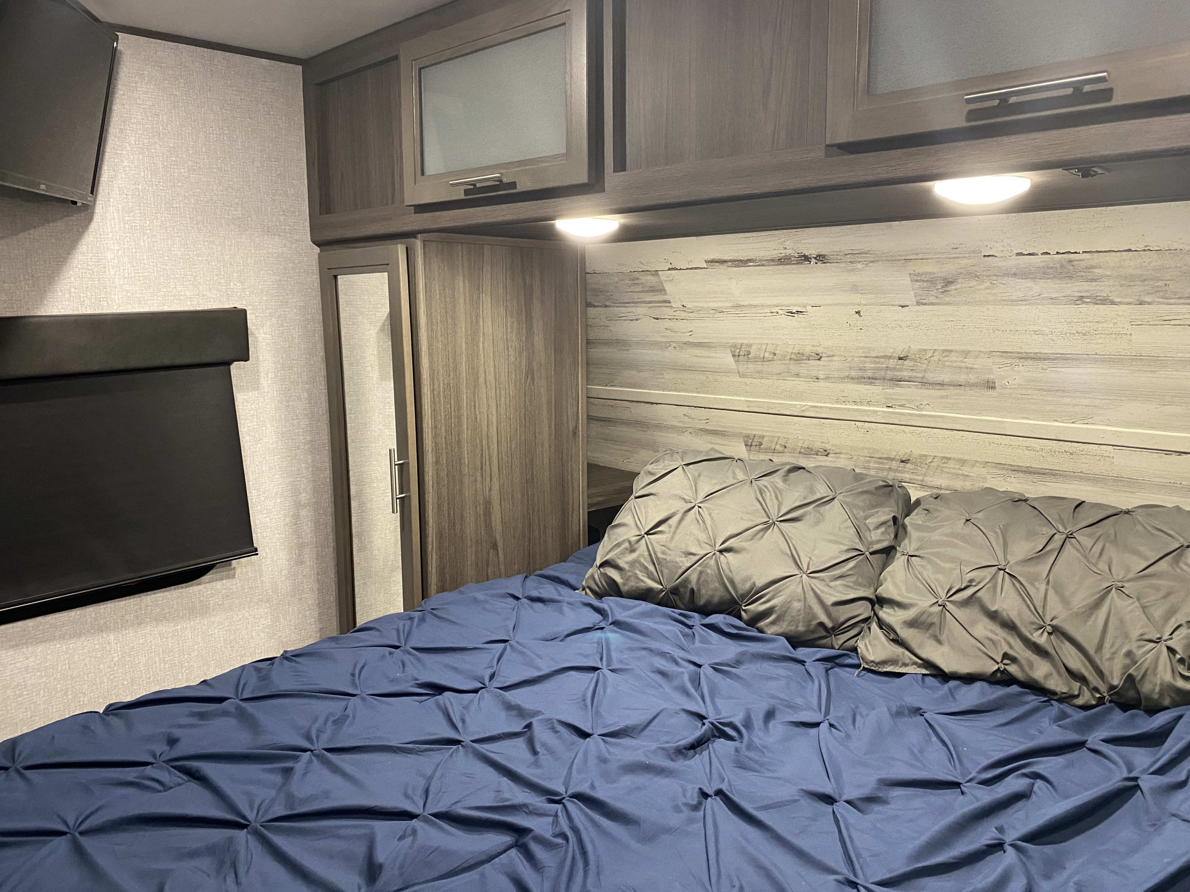 Cozy RV rental bedroom with blue bed, gray pillows, wood-look headboard, overhead storage, and wall-mounted TV.. Dutchmen Kodiak 2021
