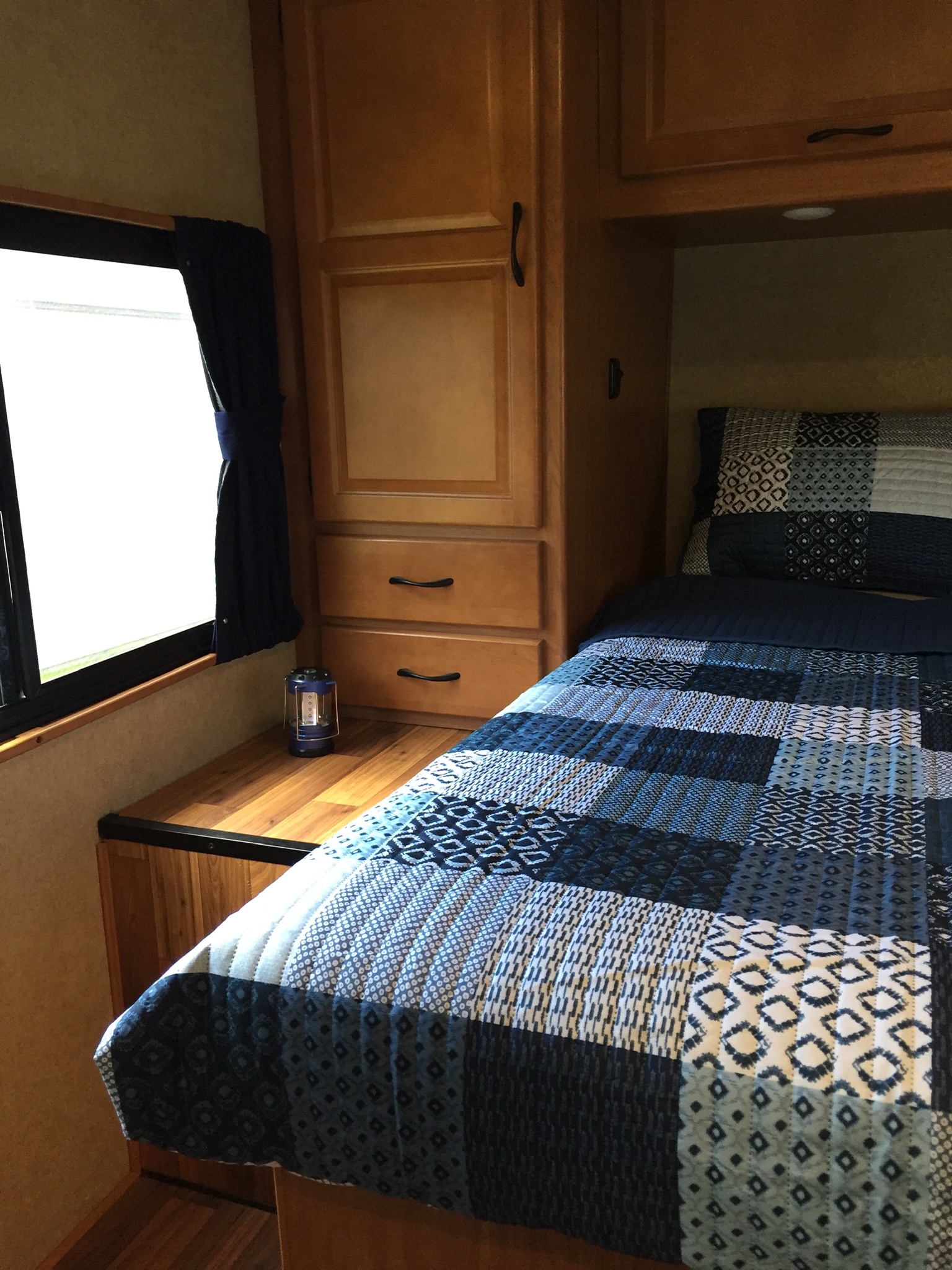 An RV rental bedroom features a comfy bed with a blue patchwork quilt, window, wood cabinetry, and a lantern.. Ford Majestic 2017