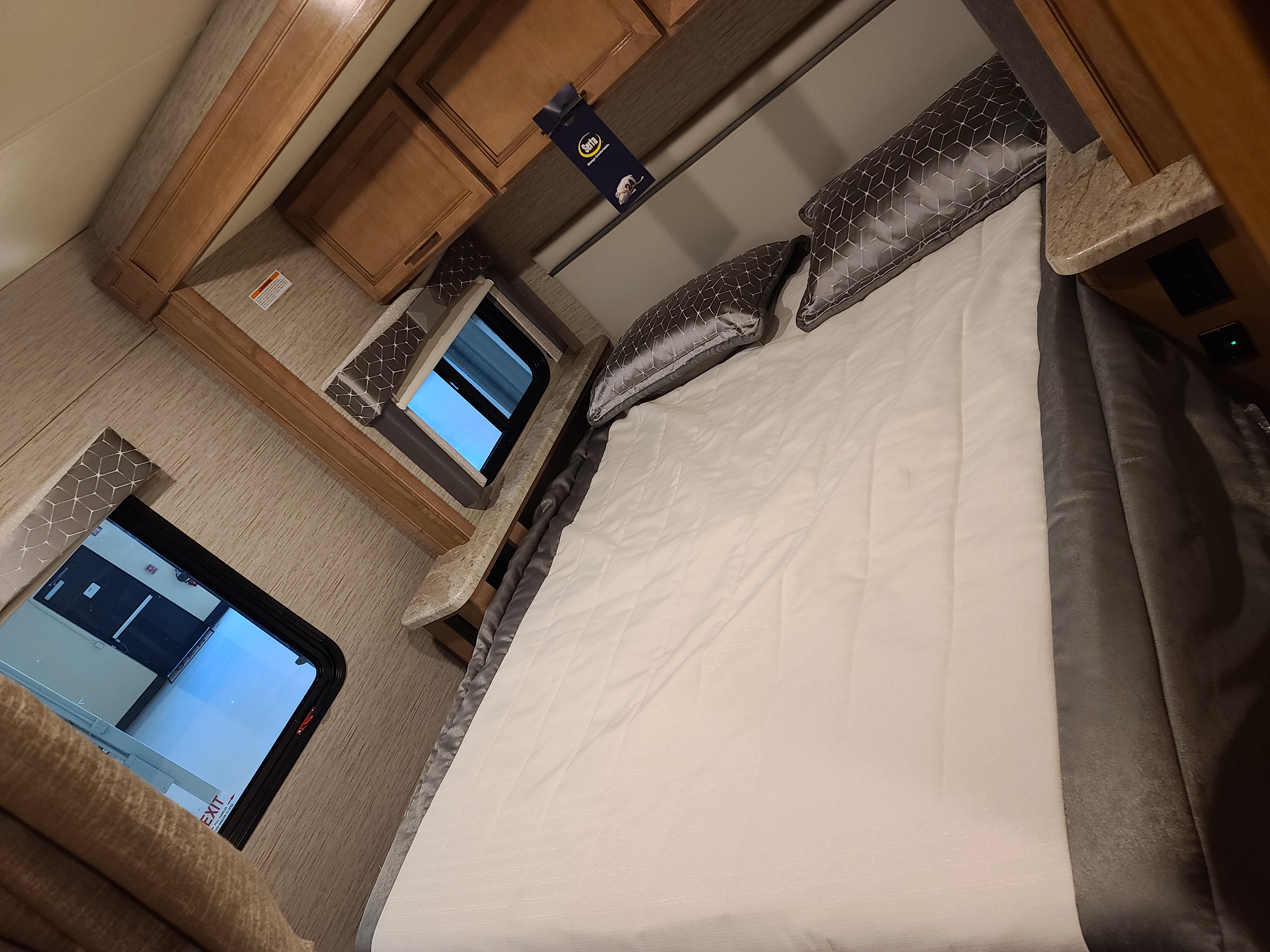 This RV rental interior showcases a cozy bedroom with a comfortable bed, grey patterned pillows, light wood cabinetry, and a window.. Thor Motor Coach Other 2021