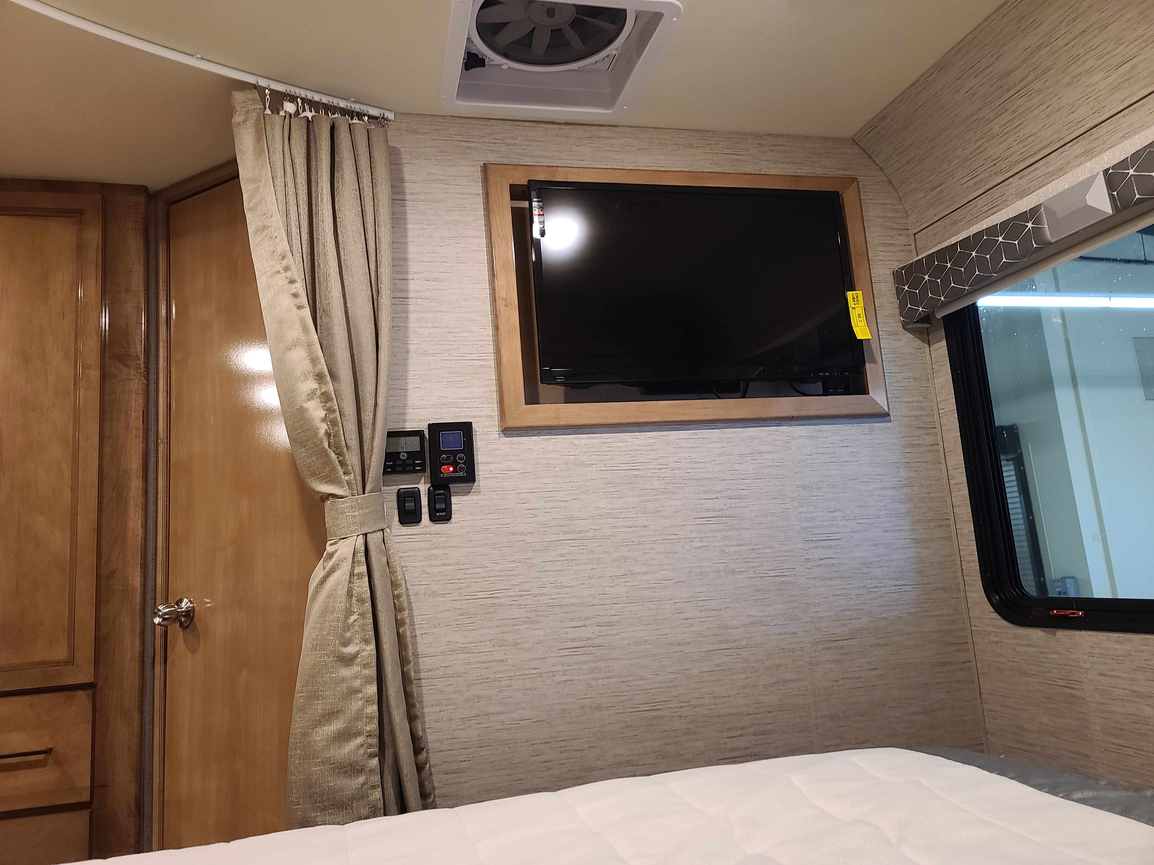 RV rental interior: bedroom with mounted TV, climate controls, and a privacy curtain by the bed and wooden cabinets.. Thor Motor Coach Other 2021