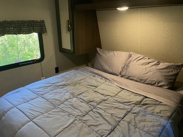 A comfortable RV rental bedroom featuring a neatly made bed with grey bedding, a window overlooking trees, and an overhead light.. Keystone Springdale 2020