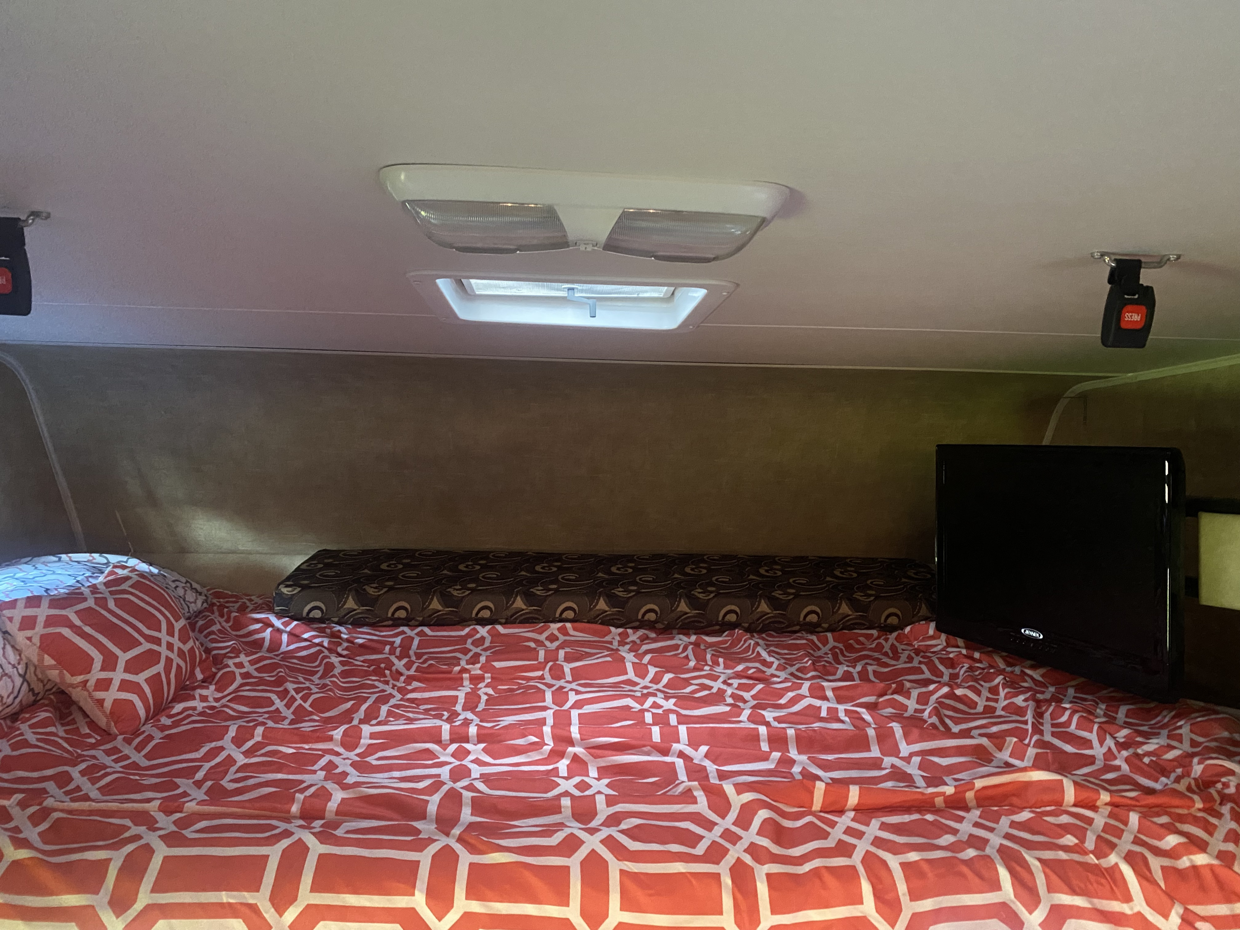 Inside an RV rental: a compact sleeping area with a vibrant red patterned bed, TV, and roof vent.. Coachmen Freelander 2014