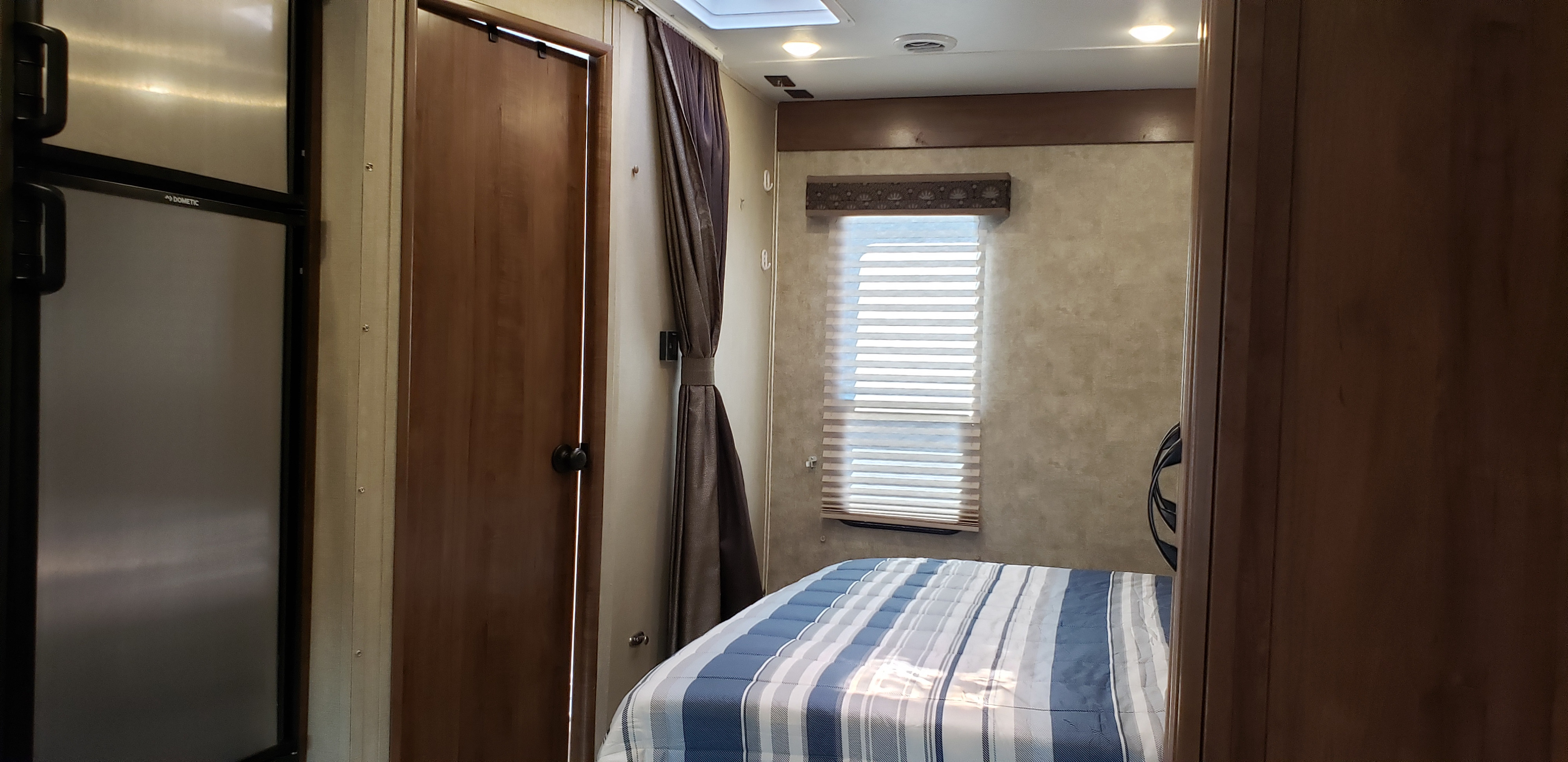 Cozy RV rental interior with compact fridge, wood door, tie-back curtain, and bedroom with blue striped bedding and window.. Gulf Stream Conquest 2018