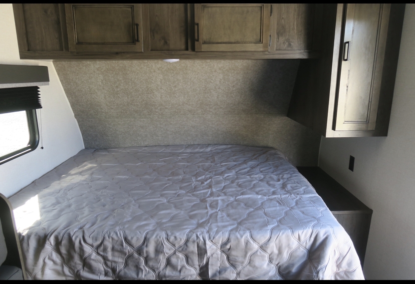 Inside an RV rental, a well-appointed bedroom features a large bed with grey quilt, overhead cabinets, and a side window.. Highland Ridge RV Open Range 2021