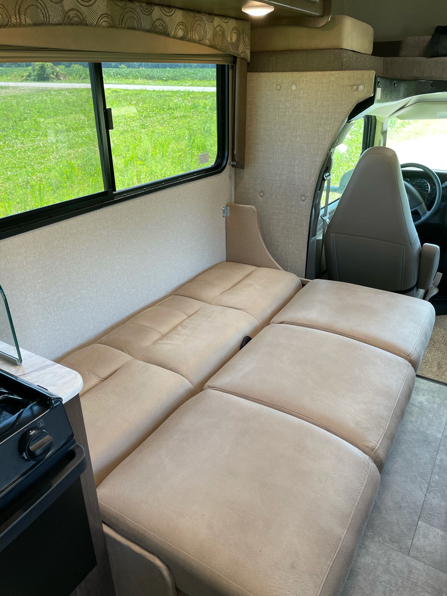 Relaxing RV rental interior with a versatile bed, large window showing green fields, and a small kitchen setup.. Coachman Freelander 2019