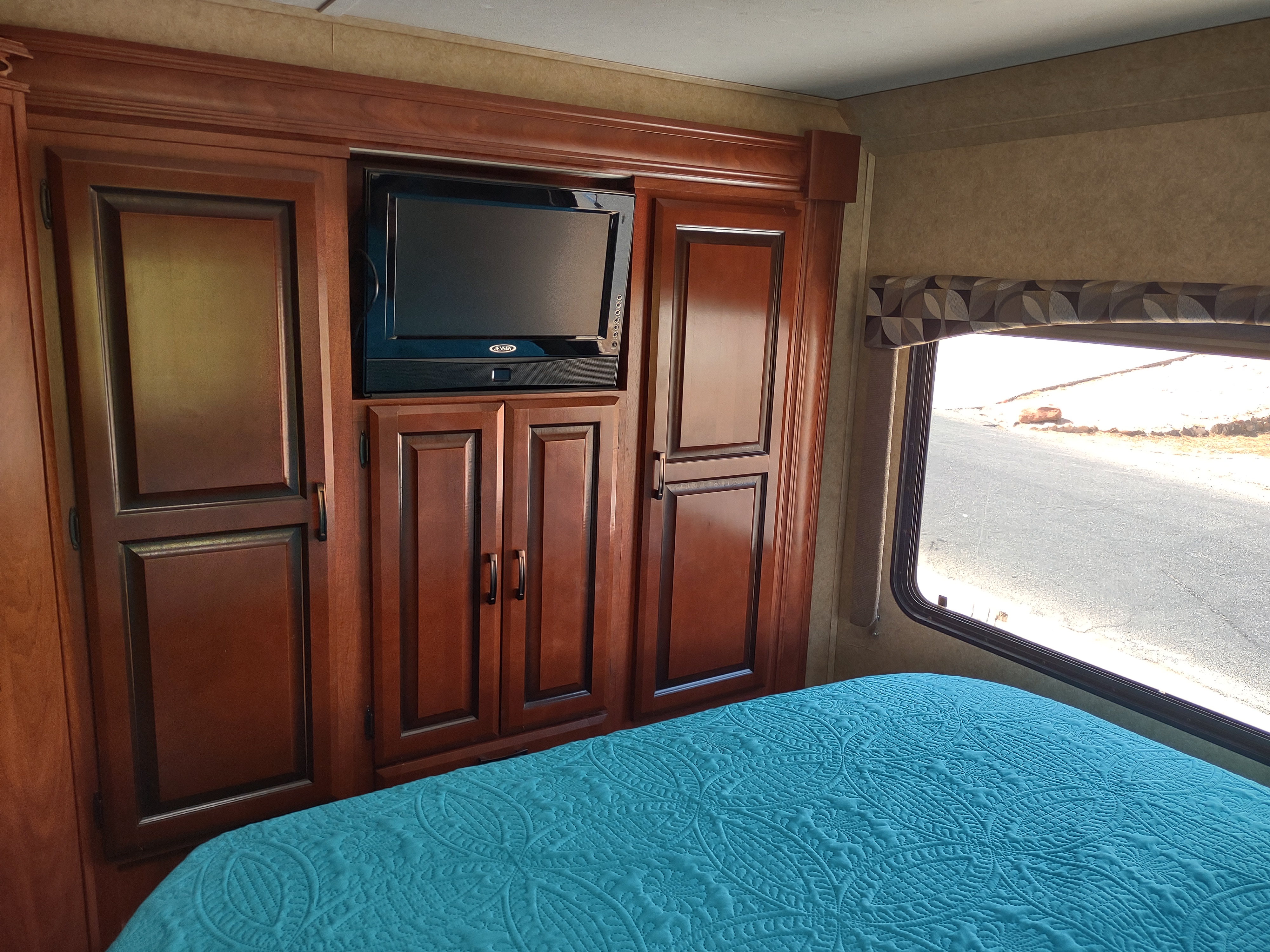 RV rental bedroom: rich wood cabinets, TV, vibrant teal quilted bed. Window shows road view outside.. Forest River Sunseeker 2012