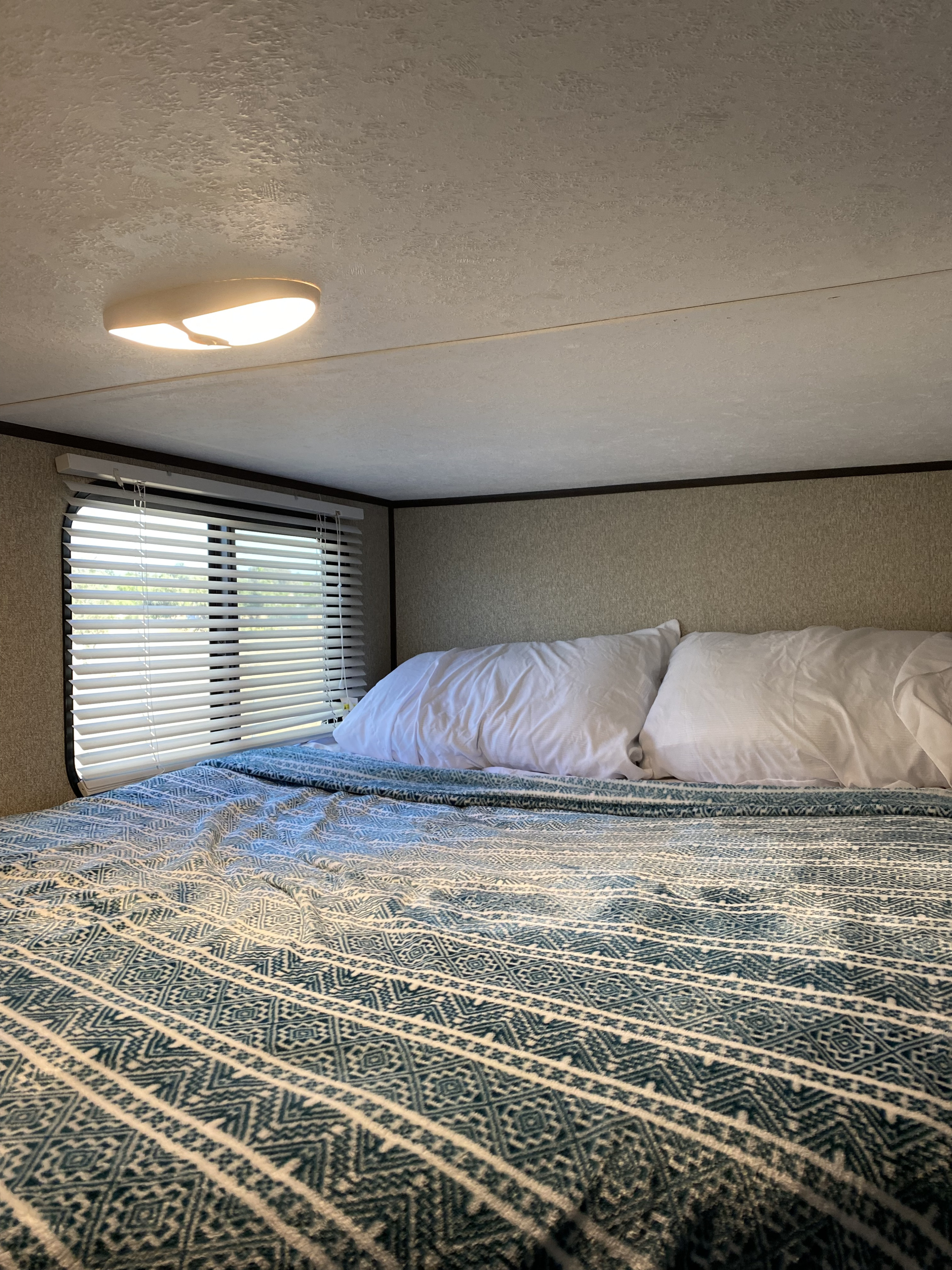 Cozy RV rental bedroom featuring a patterned blanket, white pillows, and a window with blinds, illuminated by a ceiling light.. Keystone Springdale 2018