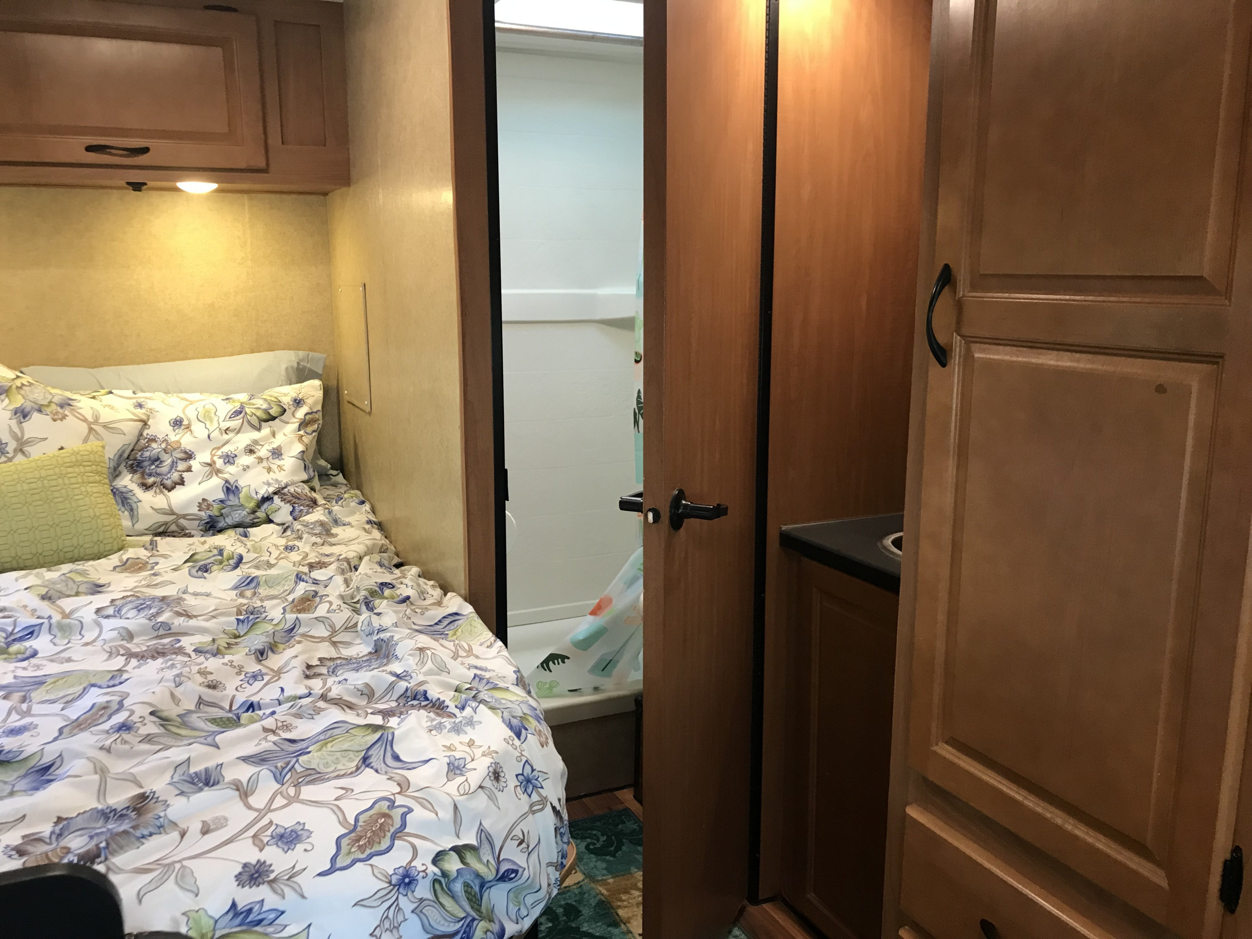RV rental interior featuring a bed with floral bedding, built-in cabinet, and peek into a shower and small kitchen space.. Thor Motor Coach Four Winds Majestic 2017