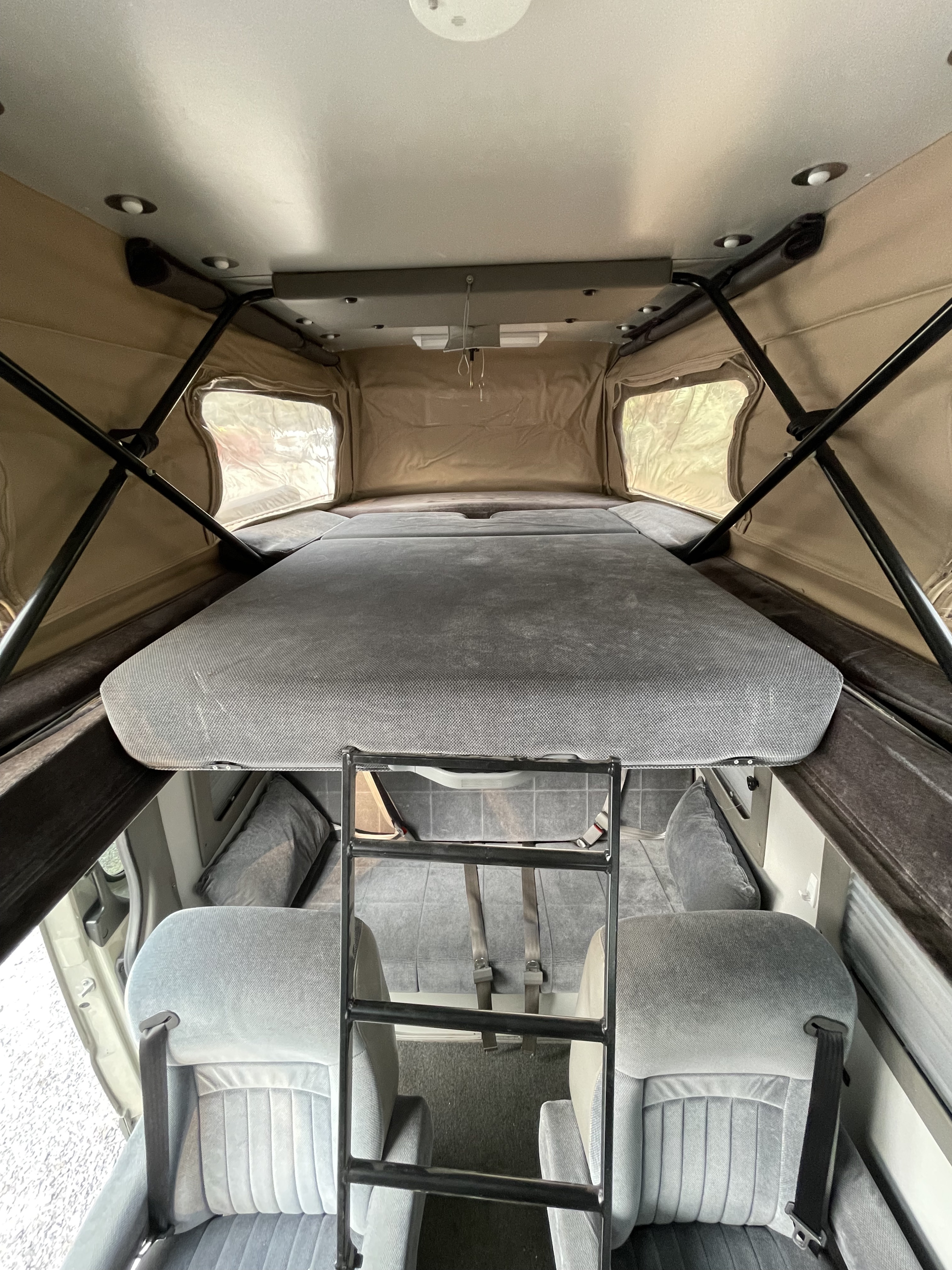 Upper sleeping berth of an RV rental camper van, accessed by a ladder. Features pop-top canvas windows and lower cabin seats.. Sportsmobile Custom Design 2013