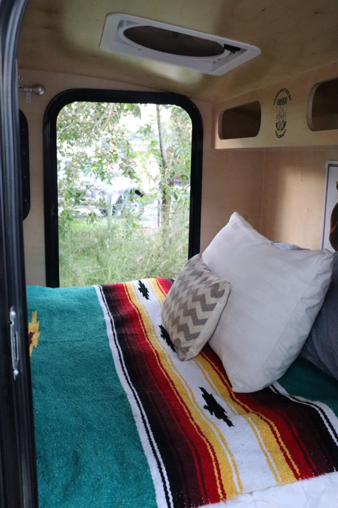 RV rental interior: cozy bed with colorful blanket and window view in a handcrafted Hiker Trailer.. Hiker Highway 2021