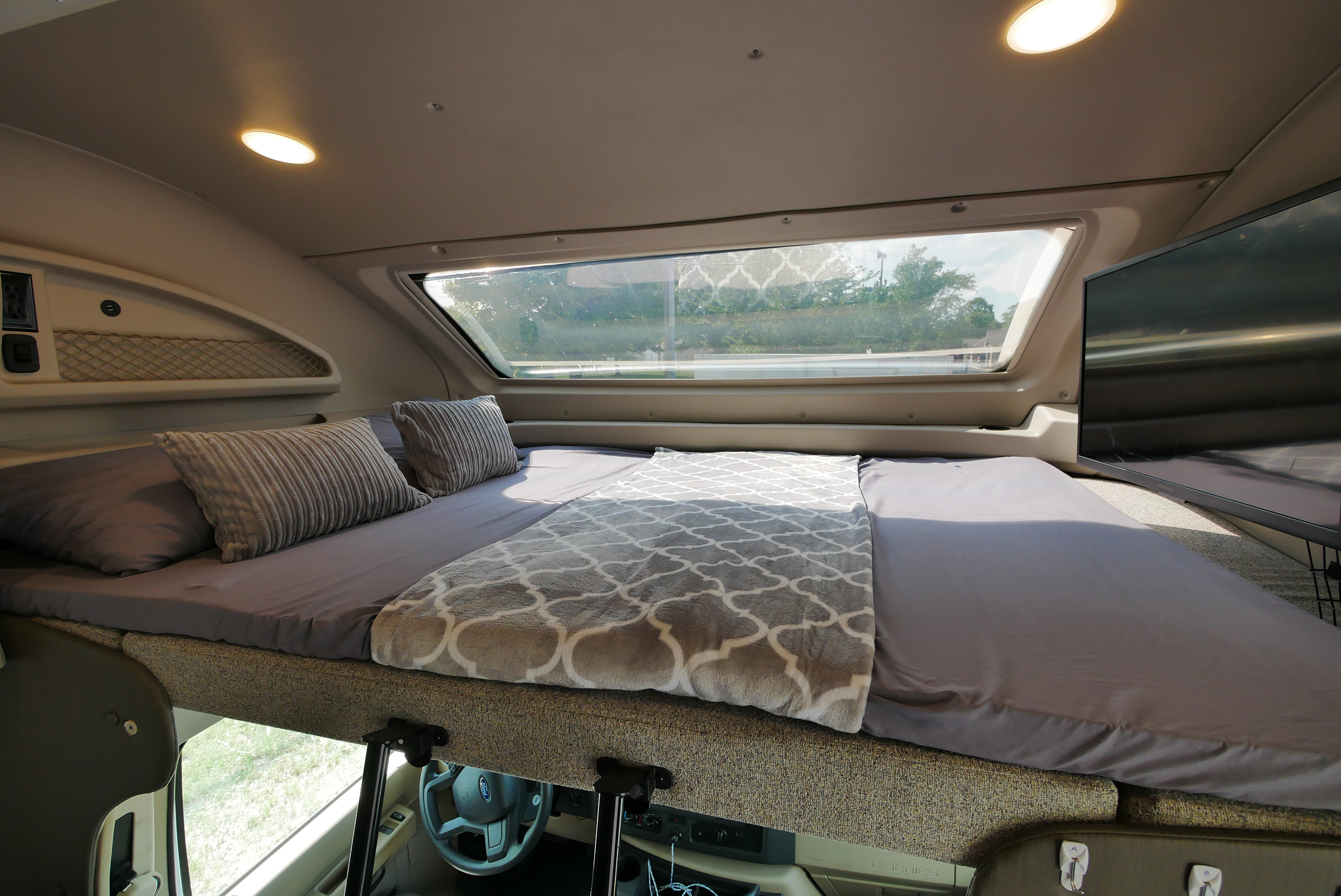 Overhead sleeping area in an RV rental, featuring a comfortable bed, large window, and TV for your travels.. Entegra Coach Other 2021