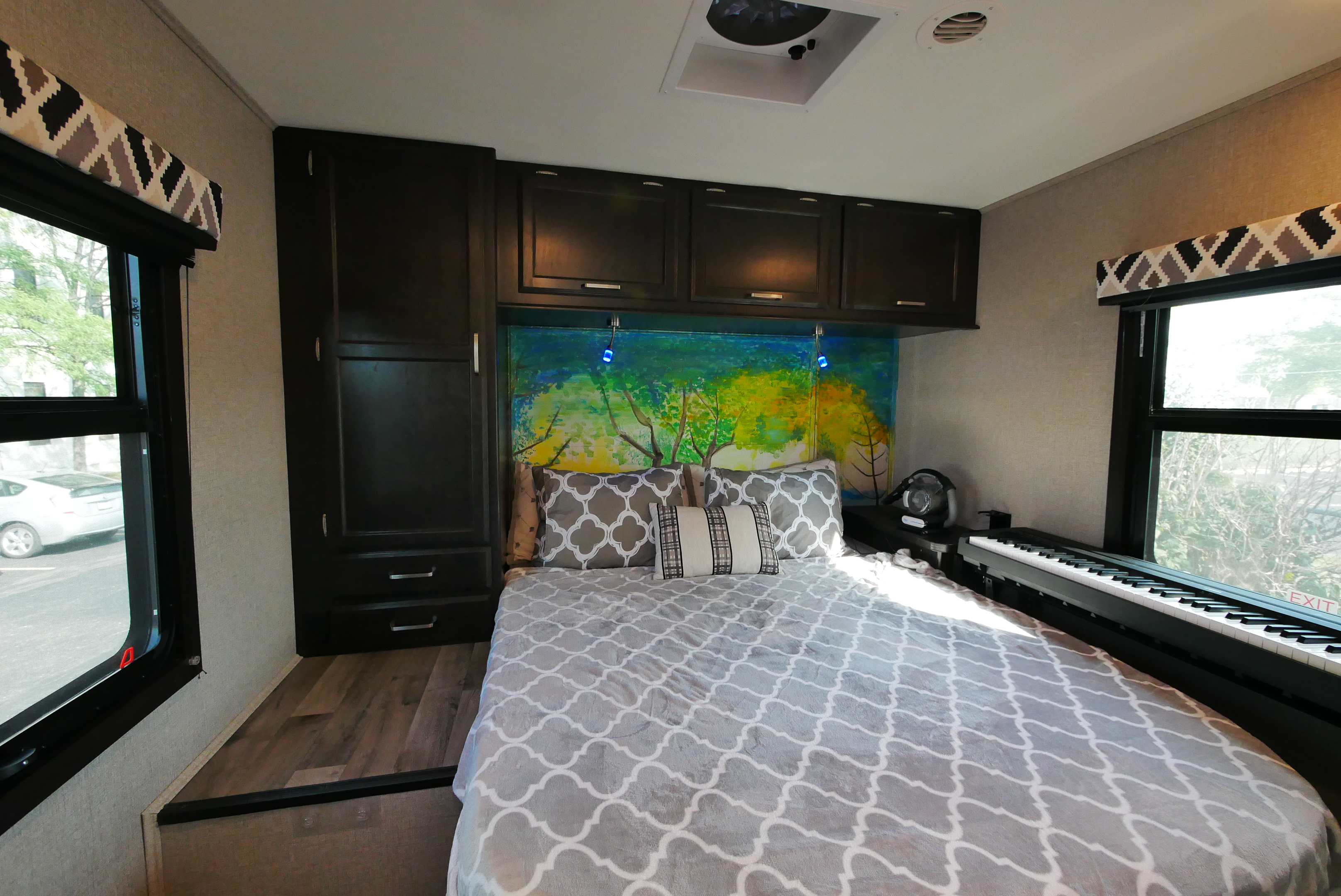 Cozy RV rental bedroom: dark cabinets, bed with grey patterned blanket, vibrant headboard mural, and a piano by the window.. Entegra Coach Other 2021