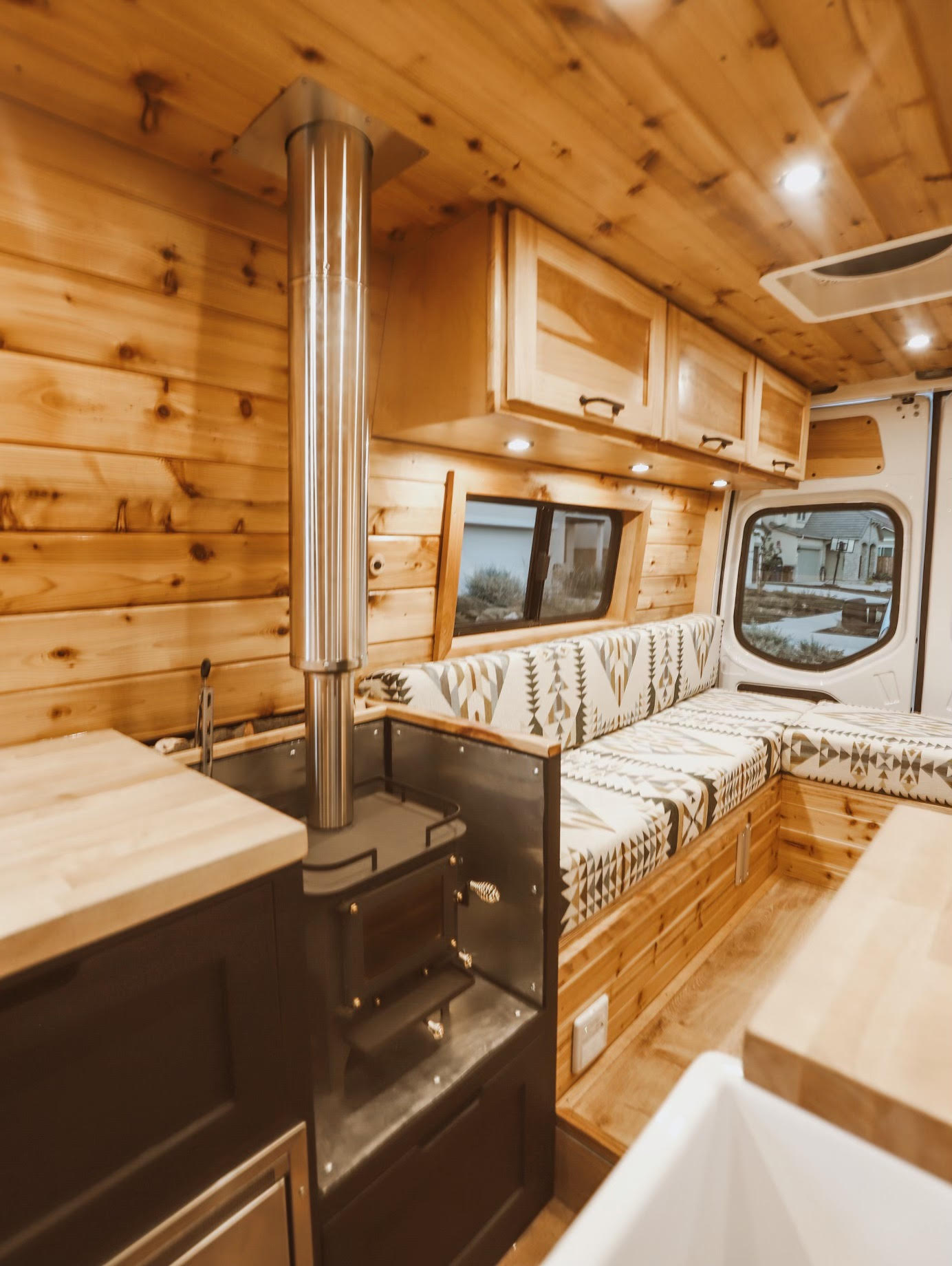 Cozy RV rental interior with a wood stove, warm wood paneling, and patterned seating, ideal for unique mobile adventures.. Other Sprinter 2019