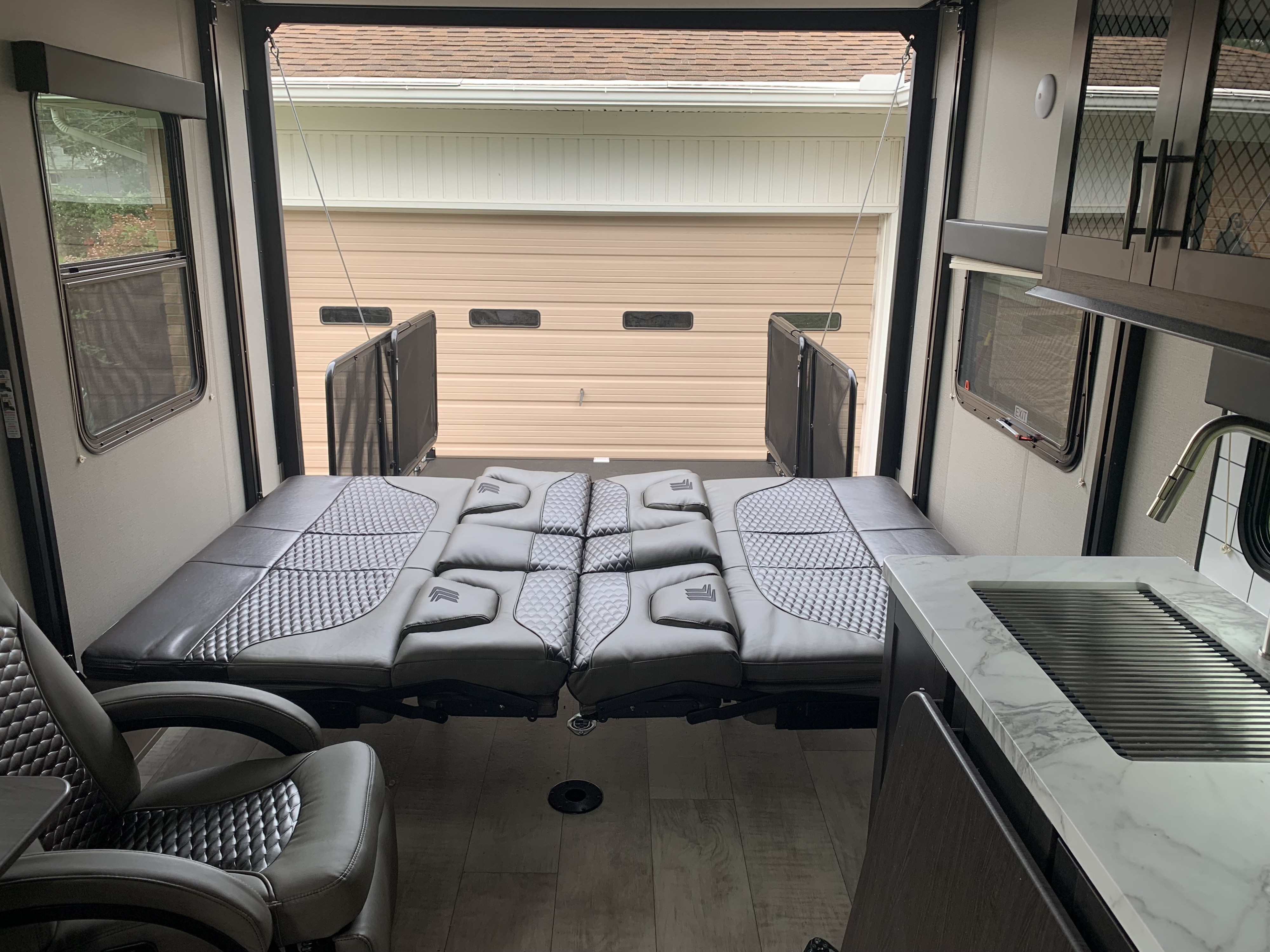 Modern RV rental interior with grey convertible bed/seating, kitchen area, and open rear ramp facing a garage.. Grand Design Momentum Toy Hauler 2022