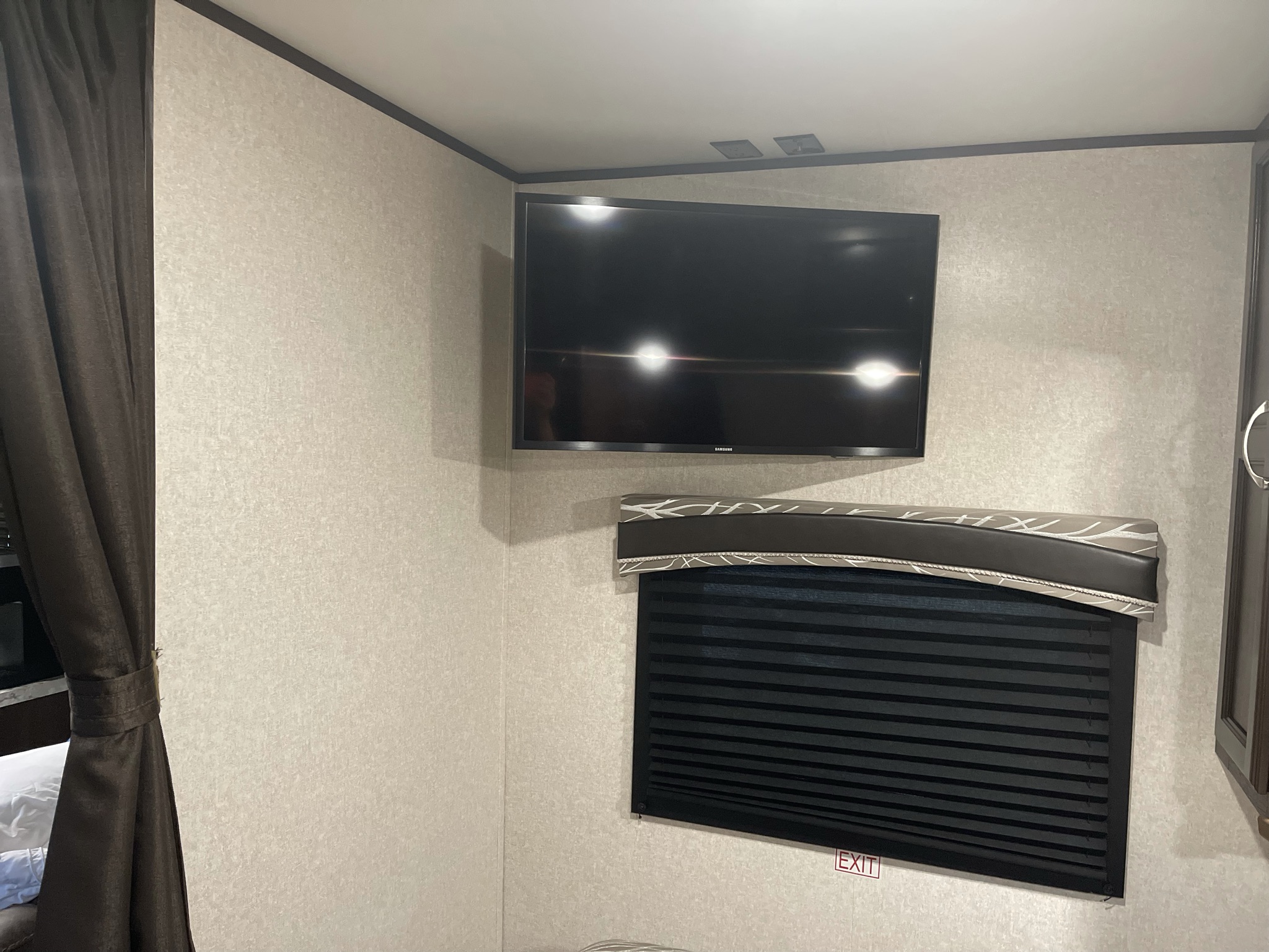 View of an **RV rental** interior, showing a wall-mounted TV on a textured wall, a black pleated blind on a window, and dark curtains.. Jayco Other 2018