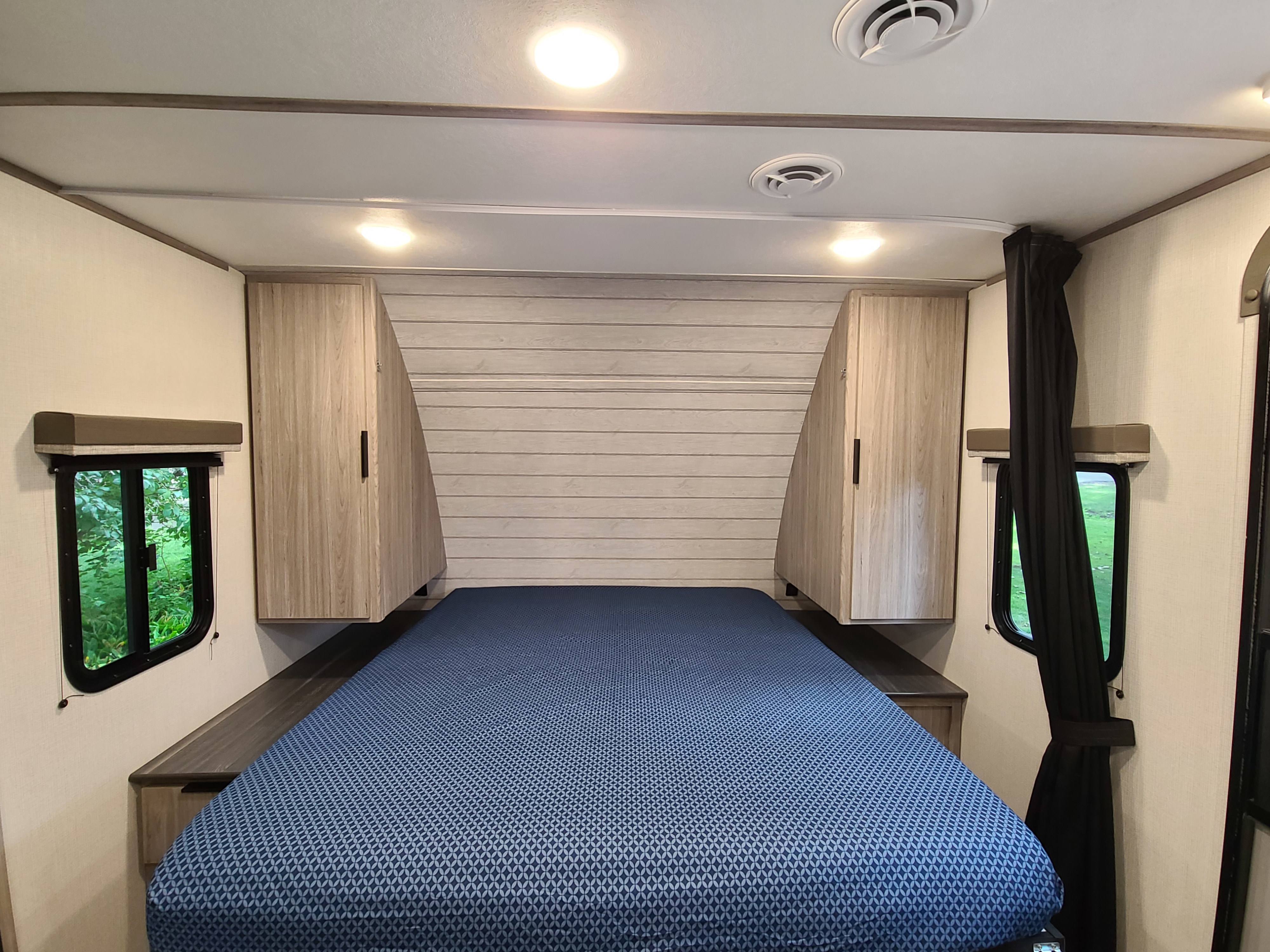 Cozy RV rental bedroom featuring a blue bed, wood-paneled headboard, and storage cabinets, with green outdoor views.. Keystone Passport 2021