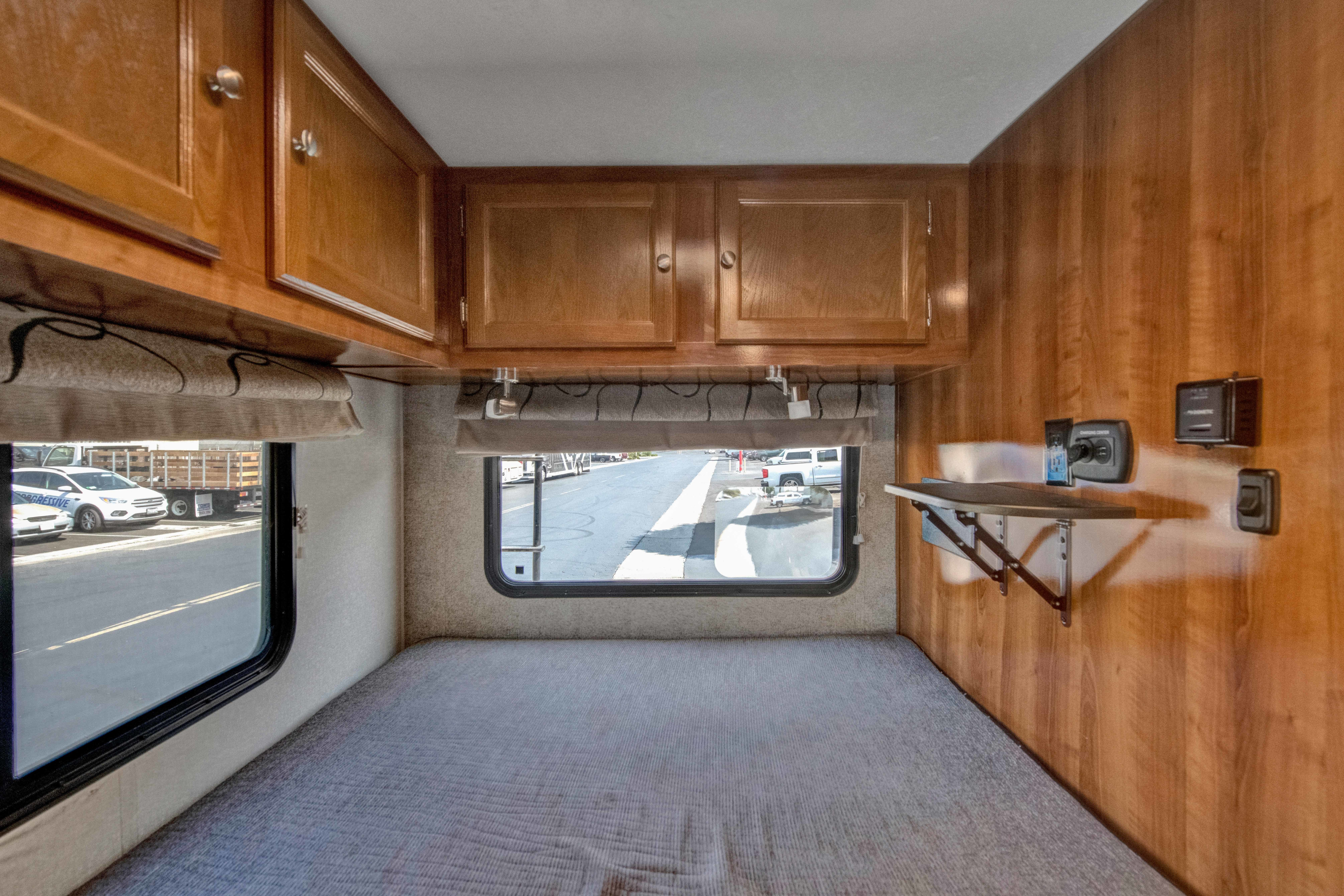 This RV rental interior features a bed, overhead wooden cabinets, a window showing a parking lot, and a fold-down shelf.. Forest River Coachmen Leprechaun 2019