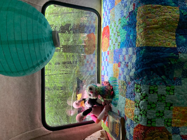 RV rental interior shows a vibrant quilt, hanging lantern, plush toys, and a lush forest view through the window.. Scamp 13' 2021