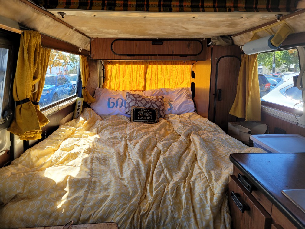 Cozy RV rental interior features a large bed with yellow bedding, yellow curtains, and an exterior view from the windows.. Volkswagen T2 Westfalia 1977