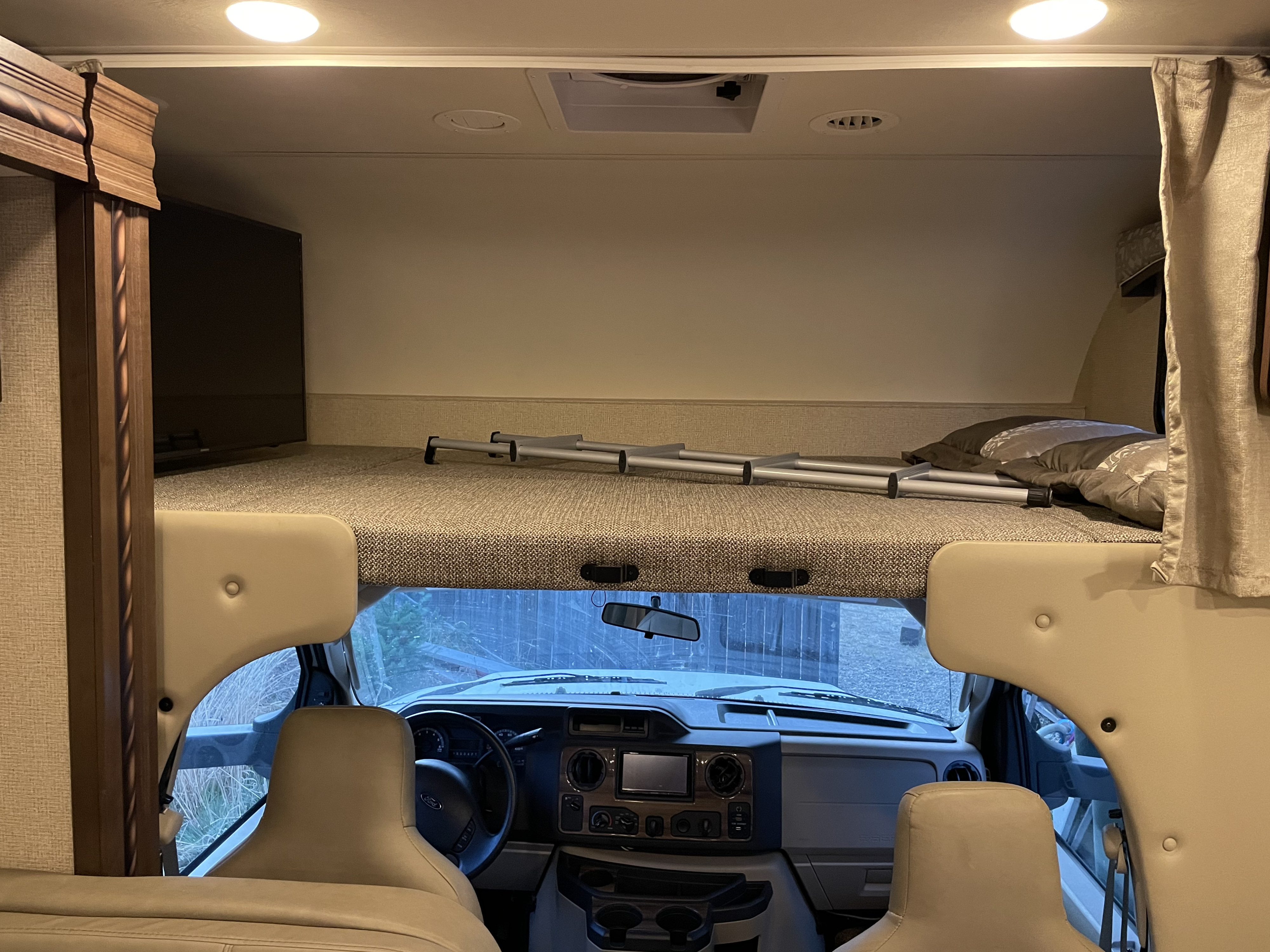 Inside an RV rental: the driver's cockpit, overhead bunk with ladder, TV, and ceiling lights/vents.. Thor Motor Coach Chateau 2017