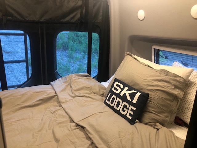Inside a comfortable RV rental: a bed with a 'SKI LODGE' pillow and views of nature outside the camper windows.. Winnebago Revel 2020