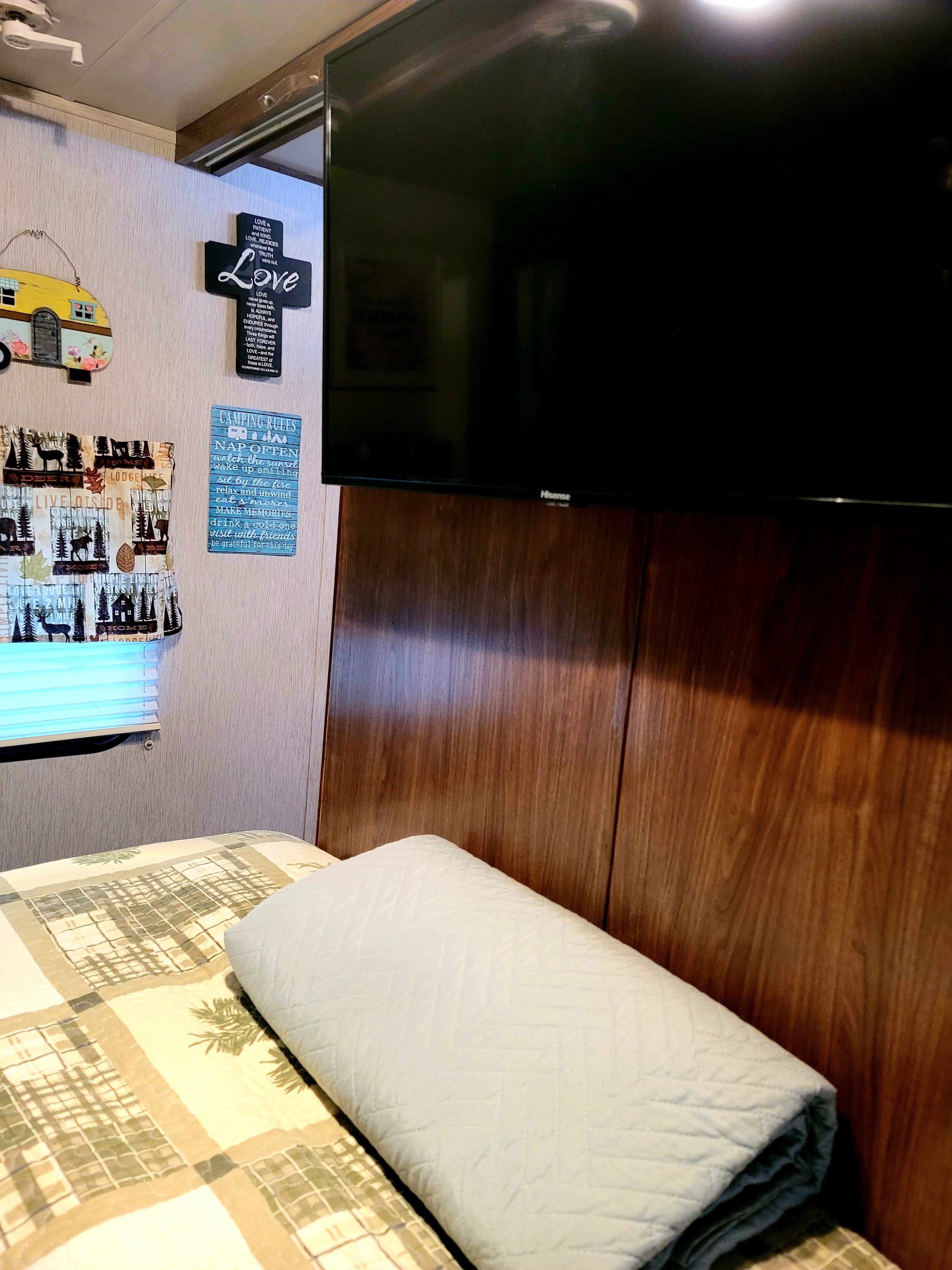 Cozy RV rental bedroom showing patterned bedding, a TV, and rustic wall decor including a 'Love' cross and 'Camping Rules' sign.. Heartland RVs Pioneer 2019